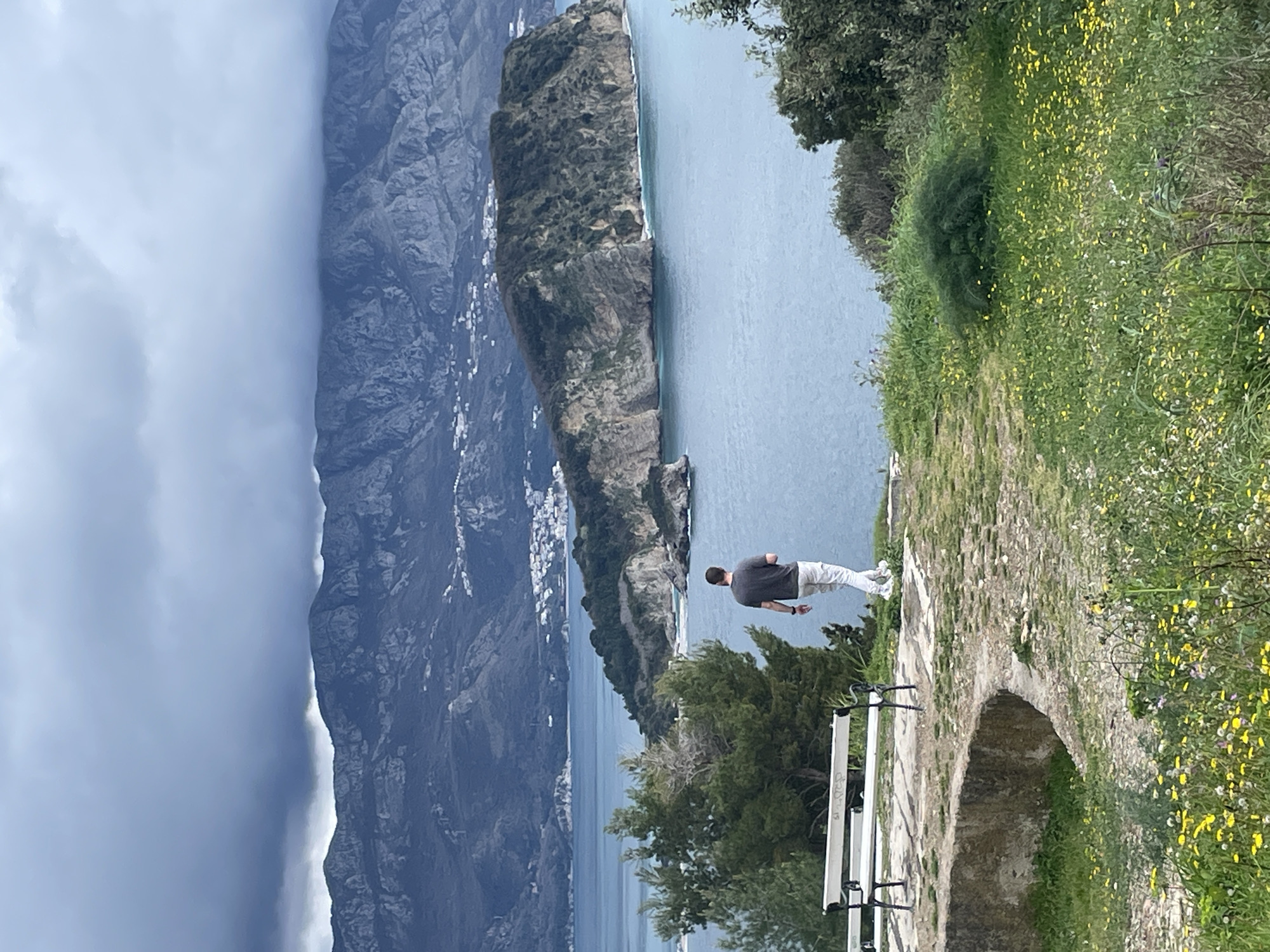 The image displays a coastal panorama from an elevated viewpoint in Budva, Montenegro, with a clear view of Sveti Nikola Island. The sky is overcast with heavy, dark grey clouds extending across the rugged, dark grey mountains in the background. Scattered white structures are visible on the lower slopes of these mountains. The Adriatic Sea in the midground is a calm, bluish-grey expanse. Sveti Nikola Island, an elongated, rocky island covered in dense green vegetation, occupies the midground, featuring steep cliffs and visible waves breaking at its base.
In the foreground, a grassy area is abundant with small yellow, white, and purple wildflowers, interspersed with patches of exposed earth. A single male subject, viewed from behind, is walking towards the edge of this scenic overlook, facing the sea and the island. He is dressed in a dark grey short-sleeved shirt, light-colored trousers, and white athletic shoes. To the left, partially obscured by a green pine tree, are two white park benches with black metal frames, positioned on a paved surface. A curved stone or concrete retaining wall is visible in the lower-left foreground.
