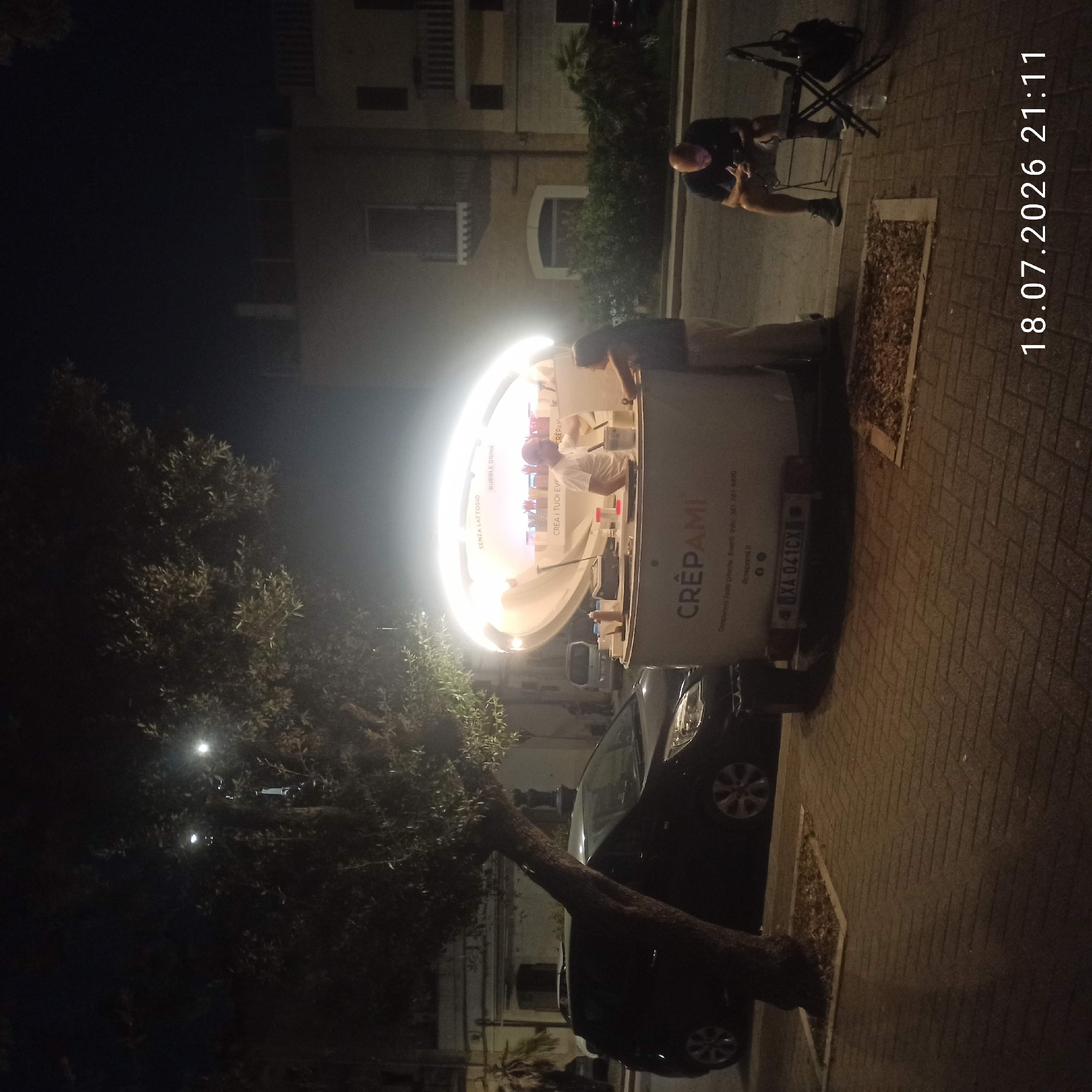 In Bitonto, Italy, at 9:11 PM on July 18, 2026, a mobile creperie named "Crepami" is illuminated by a large, oval light fixture. The creperie is white with the name "CREPAMI" prominently displayed in red lettering on its side. It appears to be set up on a sidewalk with a brick pattern. A black car is parked to the left of the creperie, and a small tree with a thick trunk stands between the car and the sidewalk.
Inside the creperie, a man in a white t-shirt is serving a customer, a woman in a dark top and light-colored pants, who is leaning over the counter. The counter is stocked with various toppings and utensils. A license plate, "XA 041CX," is visible on the back of the creperie.
To the right of the creperie, a man in a dark t-shirt and shorts is seated on a folding chair, looking at his phone. A chair and a bag are next to him. The background shows buildings with a few windows and balconies, illuminated by streetlights. The overall scene is one of a late-night food service operation in an urban setting.