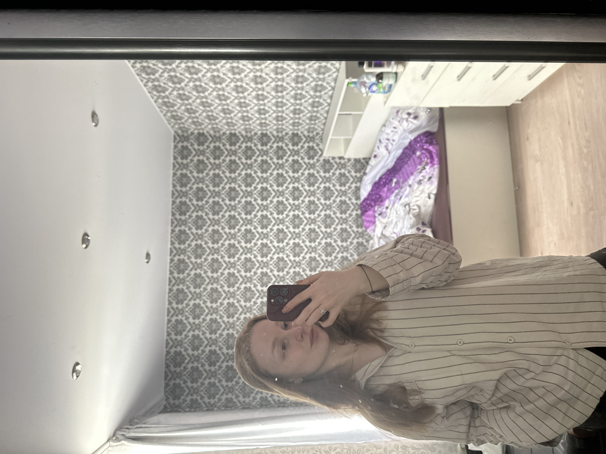 A reflection in a mirror shows a person holding a smartphone, taking a selfie. The person has fair skin and long brown hair, wearing a striped button-down shirt. The background reflected in the mirror depicts a bedroom with patterned wallpaper, a white shelf, a bed with bedding, a chest of drawers, and a wooden wall. The city of Pokrov, Russia is not discernible.
