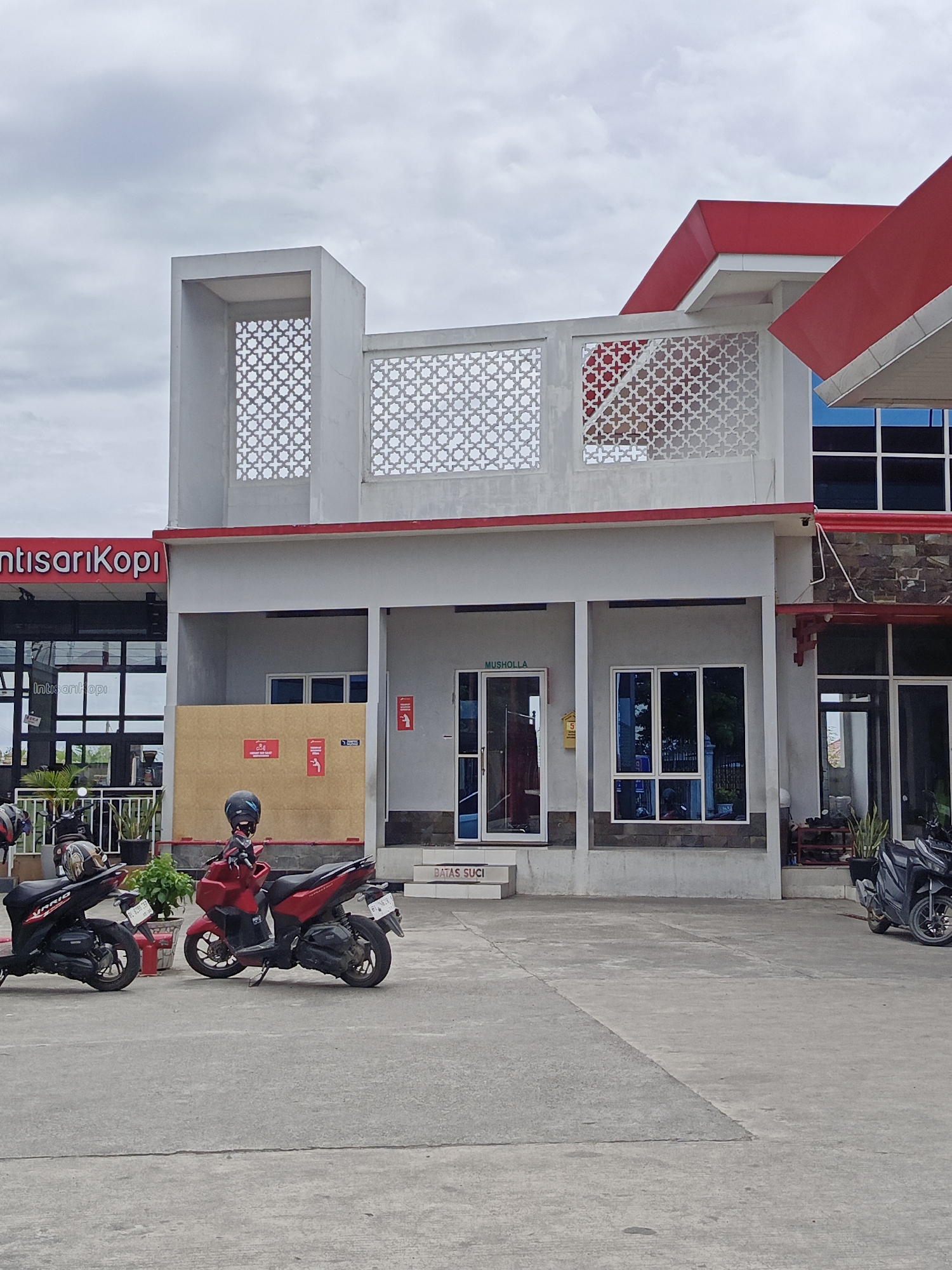 The image depicts a modern, two-story building with a red roof, likely a commercial establishment, in Cotbadatunong, Indonesia. The facade features white decorative screens with geometric patterns on the upper level. The ground floor has several entrances, including one labeled "MUSHALLA," which is a prayer room. A large, brown cardboard box is leaning against the wall near the prayer room entrance.
In front of the building, two motorcycles are parked on a concrete surface. The motorcycle on the left is black, and the one on the right is red. Both appear to be in a parked, stationary position.
The environment is an outdoor setting, possibly a parking lot or courtyard adjacent to the building. The sky is overcast with a light grey cloud cover. The building's architecture suggests a contemporary design, possibly a gas station or a rest stop given the presence of a cafe sign partially visible on the left which reads "ntisarikopi." The overall impression is one of a functional and well-maintained commercial space.
