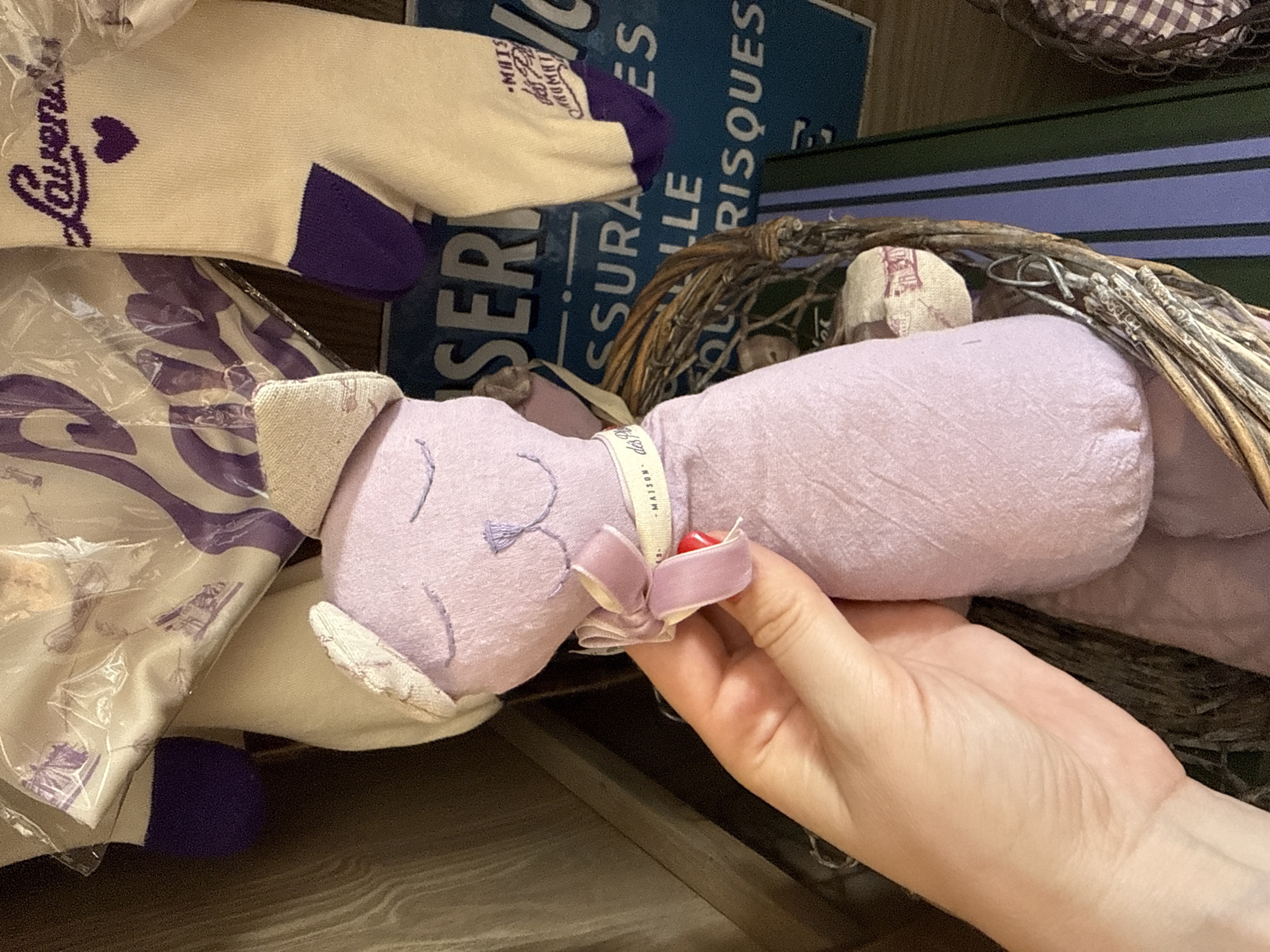 A close-up, eye-level shot shows a person's hand holding a lavender plush cat with a white bow and a small white tag that reads "-MAISON des FLEURS." The cat is positioned in front of a basket filled with dried grass. In the background, two pairs of cream-colored socks with purple trim and lettering are visible. One sock has the word "Lavenders" embroidered in purple with a small heart above it. Behind the socks, a blue sign with white lettering that appears to read "ASSURANCES" is partially visible. The lighting is natural and diffused. The image is sharp and in focus, with a shallow depth of field that blurs the background. There is no information about the location in Natukhaevskaya, Russia.