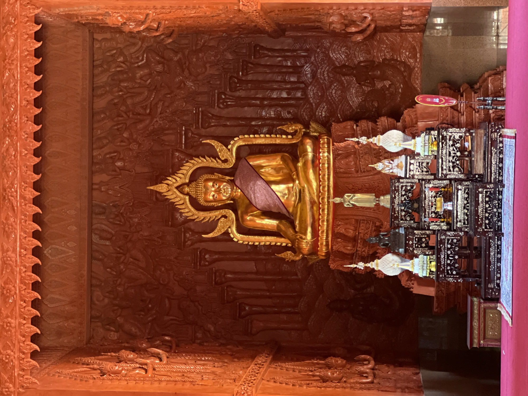The image depicts the interior of a Buddhist temple, likely located in Baan Hua Thanon, Thailand. The primary subject is a large, gilded statue of Buddha seated on a raised platform. The statue is adorned with a purple sash. The background is intricately carved with religious imagery in a reddish-orange hue, featuring figures and architectural elements. In front of the Buddha statue, there are several smaller decorative objects and altars, including miniature Buddha statues, ornate stands with intricate designs, and candles. The scene is illuminated by natural light, casting shadows and highlighting the textures of the carvings and the golden Buddha. The overall impression is one of reverence and artistic craftsmanship characteristic of Thai Buddhist temples.