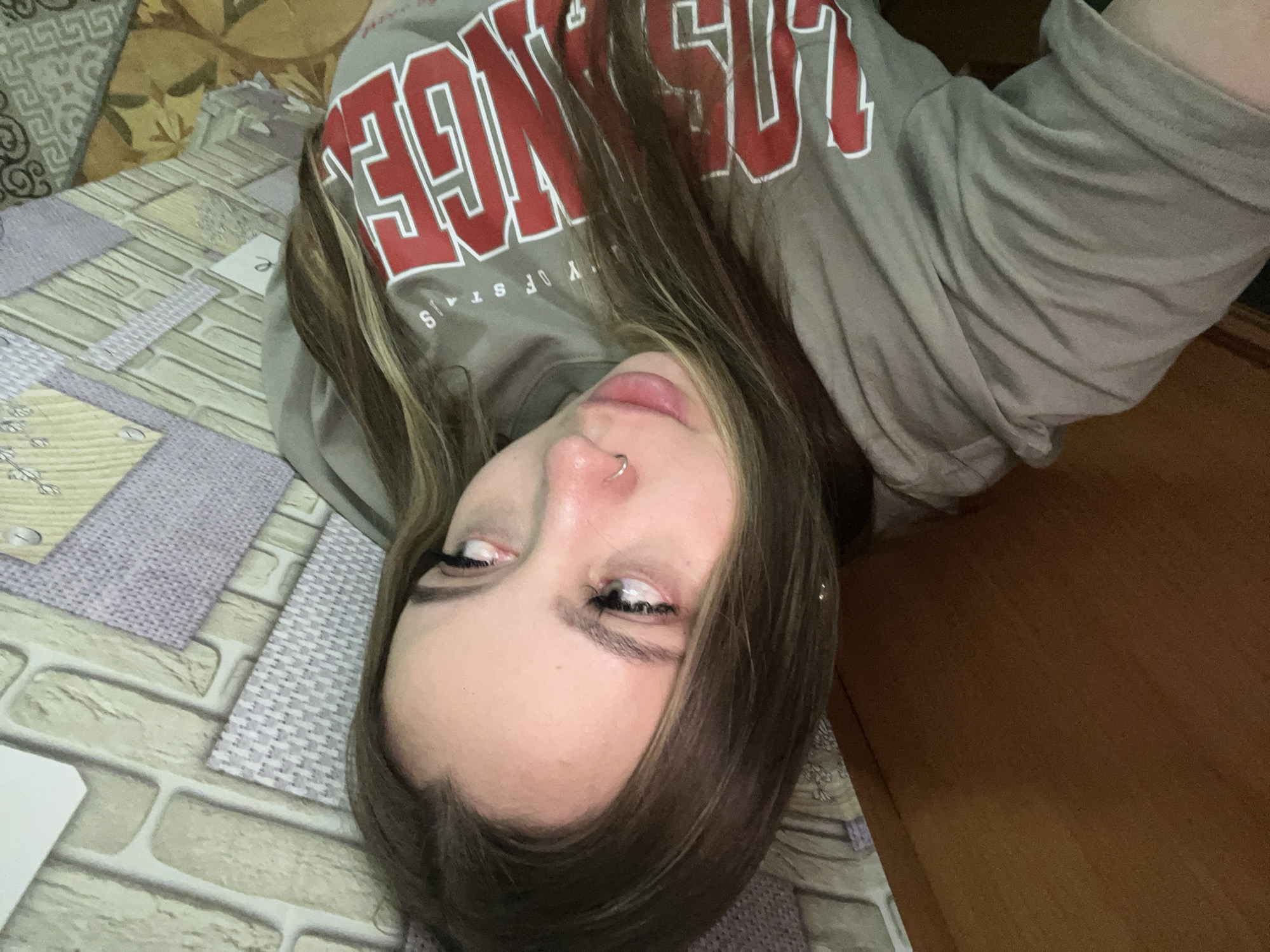 This media file features a young individual, likely female, lying down and taking a selfie indoors. The subject has long, straight brown hair with lighter streaks, framing her face. She is wearing a casual, long-sleeved gray sweatshirt. Prominently displayed on the shirt are large red block letters that appear to spell "LOS ANGELES," with smaller, partially visible text below it, possibly "UNIVERSITY OF STATES" or a similar phrase. A small silver hoop nose piercing is visible on her right nostril. Her eyes are open, looking towards the camera, with visible eyeliner and mascara. Her lips are slightly parted in a relaxed expression.
The background reveals a textured indoor setting, likely a bed or sofa. Various patterned fabrics or textiles are visible, including a light-colored pattern resembling bricks, a section of light purple-grey woven fabric, and another area with a floral or geometric design in beige and brown tones. To the right of the frame, a plain brown wooden surface is discernible. The lighting suggests an indoor environment, possibly during the daytime. The overall tone is casual and personal. The context provided indicates the photo was taken in Port-Katon, Russia.