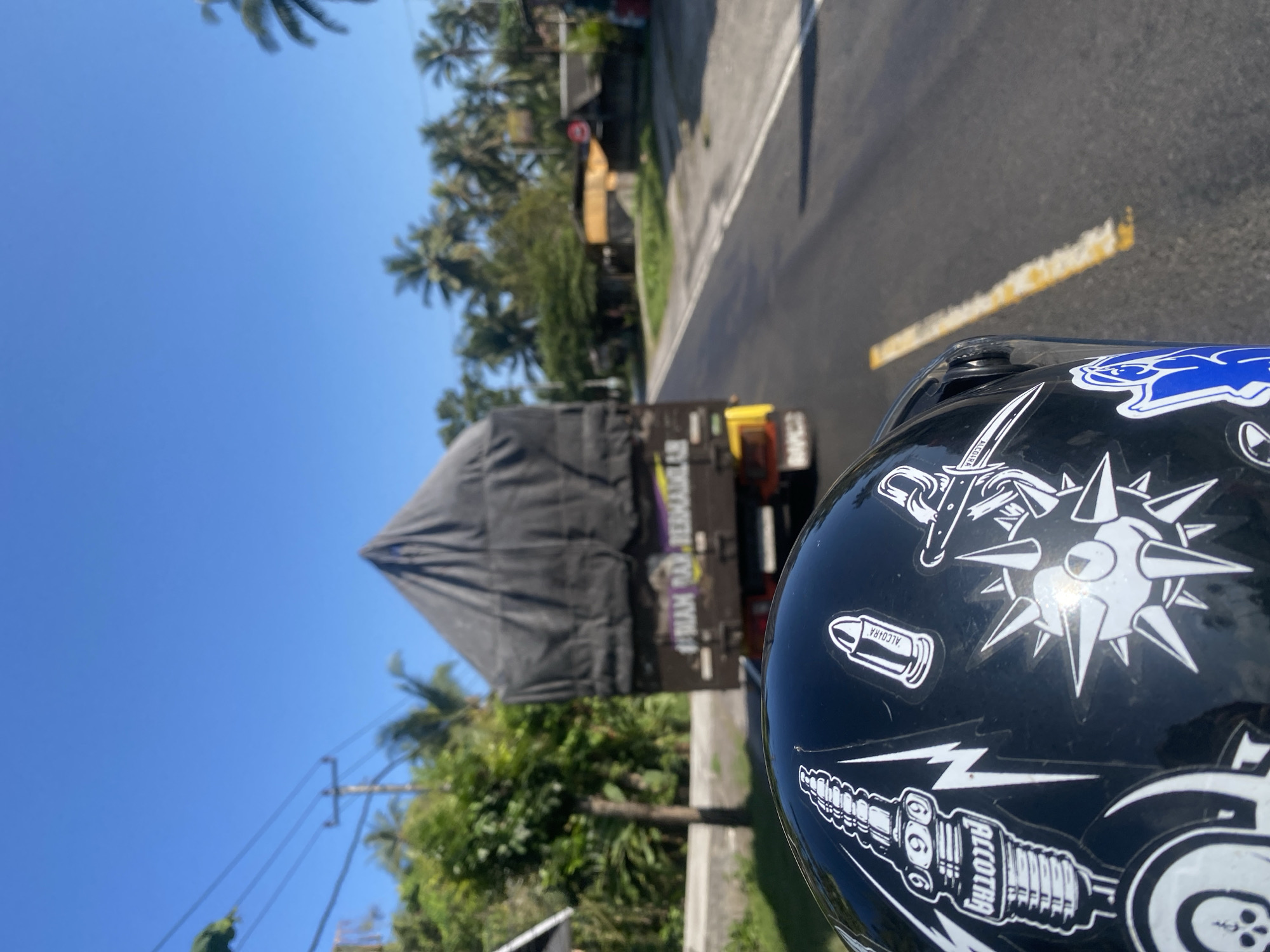 In Selemadeg Barat, Indonesia, a black motorcycle helmet, adorned with various white stickers including a spark plug, a bullet, a skull, and a spiky star, is in the foreground. The helmet is facing away from the viewer, implying a rider is present, though they are not visible. Behind the helmet, a black tarp covers the top of a truck that is traveling on a paved road. The truck has yellow accents on its rear and signage that reads "#IRIAM DAM BERMAKMUR". The road is lined with lush green vegetation, including palm trees, and power lines are visible in the distance. The sky is a clear, bright blue. The scene suggests a daytime journey on a sunny day in a tropical environment.