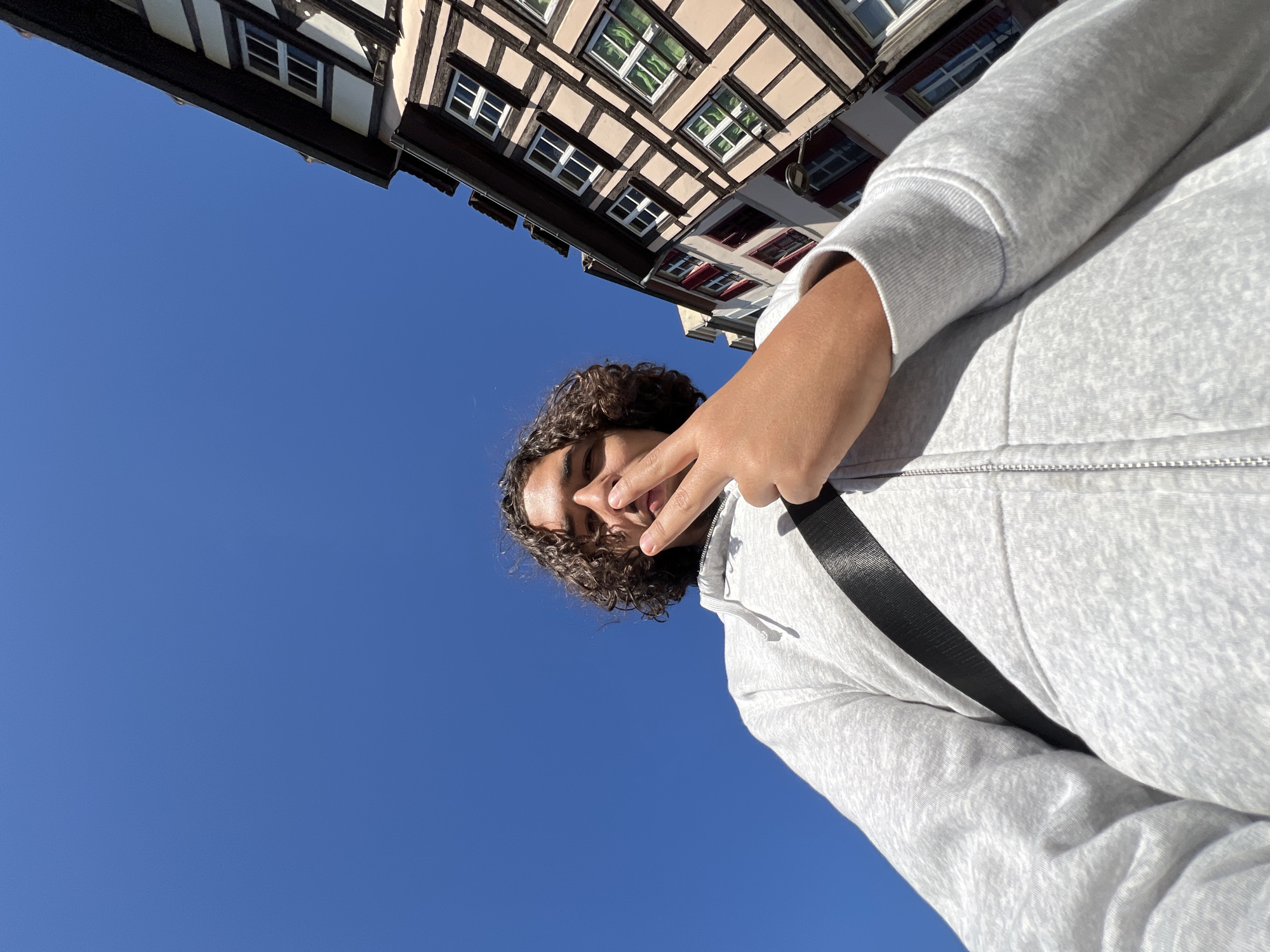 A low-angle photograph shows a person with curly dark hair wearing a light gray zip-up hoodie and a black shoulder strap against a clear blue sky. The person is holding up their hand in a peace sign. In the upper left corner of the image, portions of a traditional half-timbered building are visible. The building's facade features white-framed windows and dark brown wooden beams. This architectural style is characteristic of the Alsace region. Given the architectural style, the location is most likely somewhere in the city of Strasbourg, France.