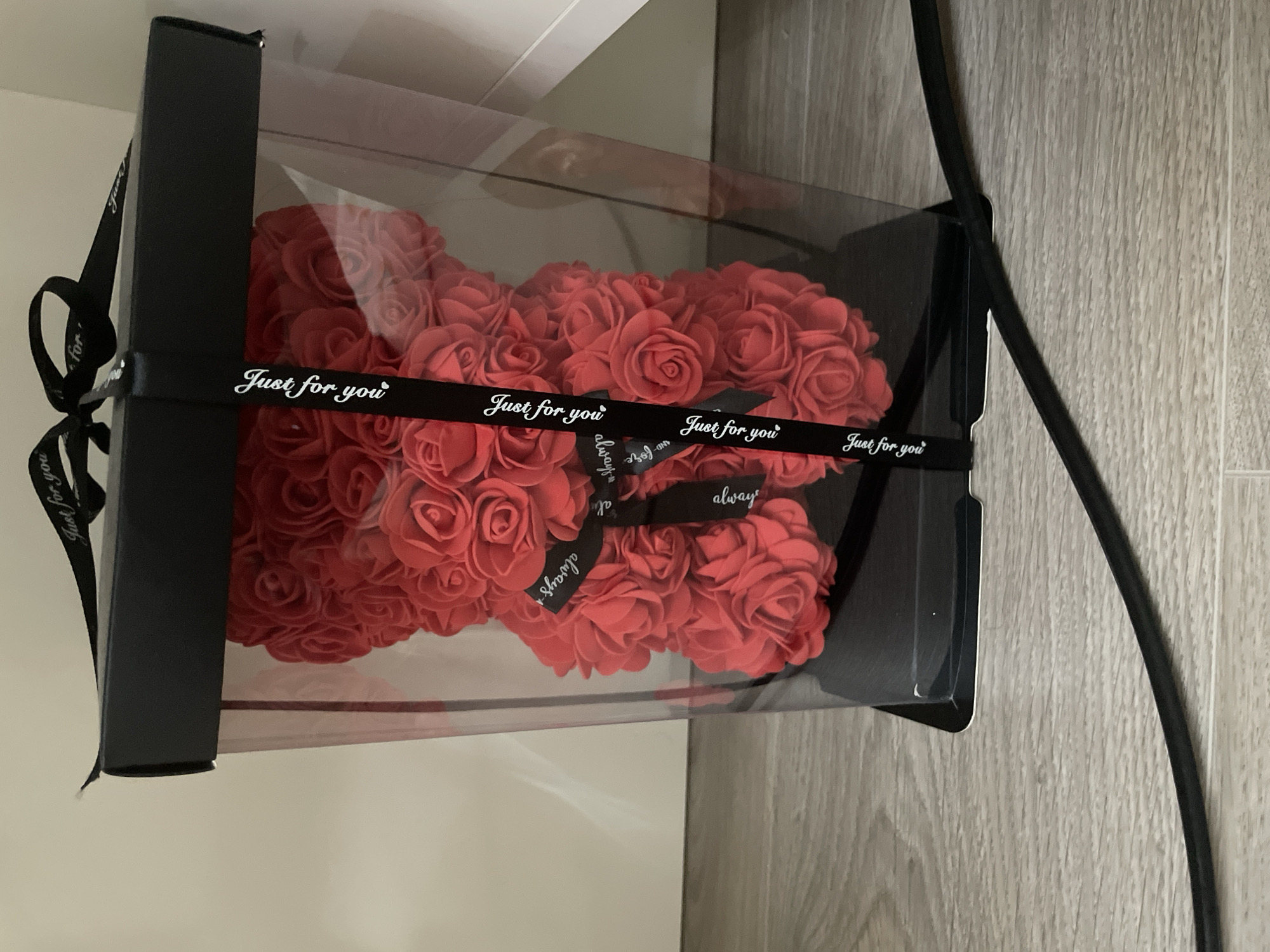 The image displays a red rose bear, a teddy bear-shaped ornament crafted from numerous artificial red roses, enclosed within a clear plastic gift box with black cardboard sides. A black ribbon featuring white text is tied around the box and the rose bear. The ribbon prominently displays the phrases "Just for you" and "always" in a decorative white font. The rose bear is composed of densely packed, individually sculpted rose blooms, creating a textured surface.
The scene is set indoors, with the gift box resting on a light-colored wooden surface, likely a floor or table, and a light-colored wall or vertical surface visible in the background. A black electrical cable is visible curving along the right side of the frame, partially obscuring the wooden background. The lighting is even, typical of an indoor setting. The clear packaging and decorative ribbon suggest that the item is a gift, intended to convey affection or celebrate an occasion, in Ihtiman, Bulgaria. No people are visible, and no specific activity is taking place other than the presentation of the object.