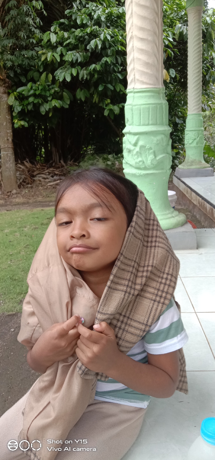 The image displays a young child, appearing female, positioned outdoors in Ranugedang, Indonesia. The child, with dark hair and a light complexion, looks directly forward with a neutral expression, exhibiting a slight pout. She wears a striped white and light green short-sleeved shirt under a light brown, possibly tan, jacket or hooded garment. A brown and black plaid fabric is draped over her head and shoulders, functioning as a hood or wrap. Her hands are clasped near her chest, holding a small, pink, possibly iridescent decorative object. To the child's right, multiple decorative pillars, fluted and carved with ornamental designs, are visible; they are painted white with light green bases. These pillars rest on a light-colored tiled surface. The background on the left and center consists of dense green foliage, including various trees and bushes. A tree trunk and piles of dried branches or wood are discernible on the far left. A patch of green grass and a dark ground surface are visible in the lower left. A blue plastic cap is partially visible in the lower right corner of the frame.