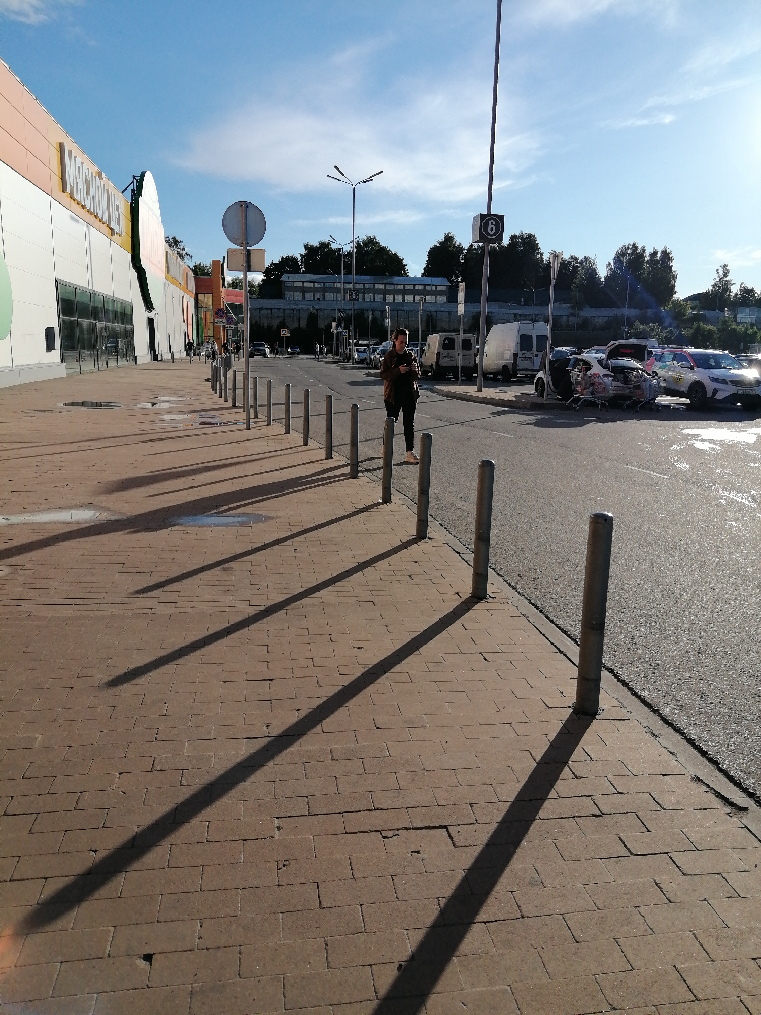 The image shows a woman standing on a brick sidewalk, looking at her phone. She is wearing a brown jacket and dark pants. The sidewalk is next to a paved road with cars parked on the side. There are several bollards along the sidewalk. In the background, there is a large building with signs, trees, and a blue sky with some clouds. The sun is shining, casting long shadows on the sidewalk. The scene appears to be a sunny day in a commercial area.