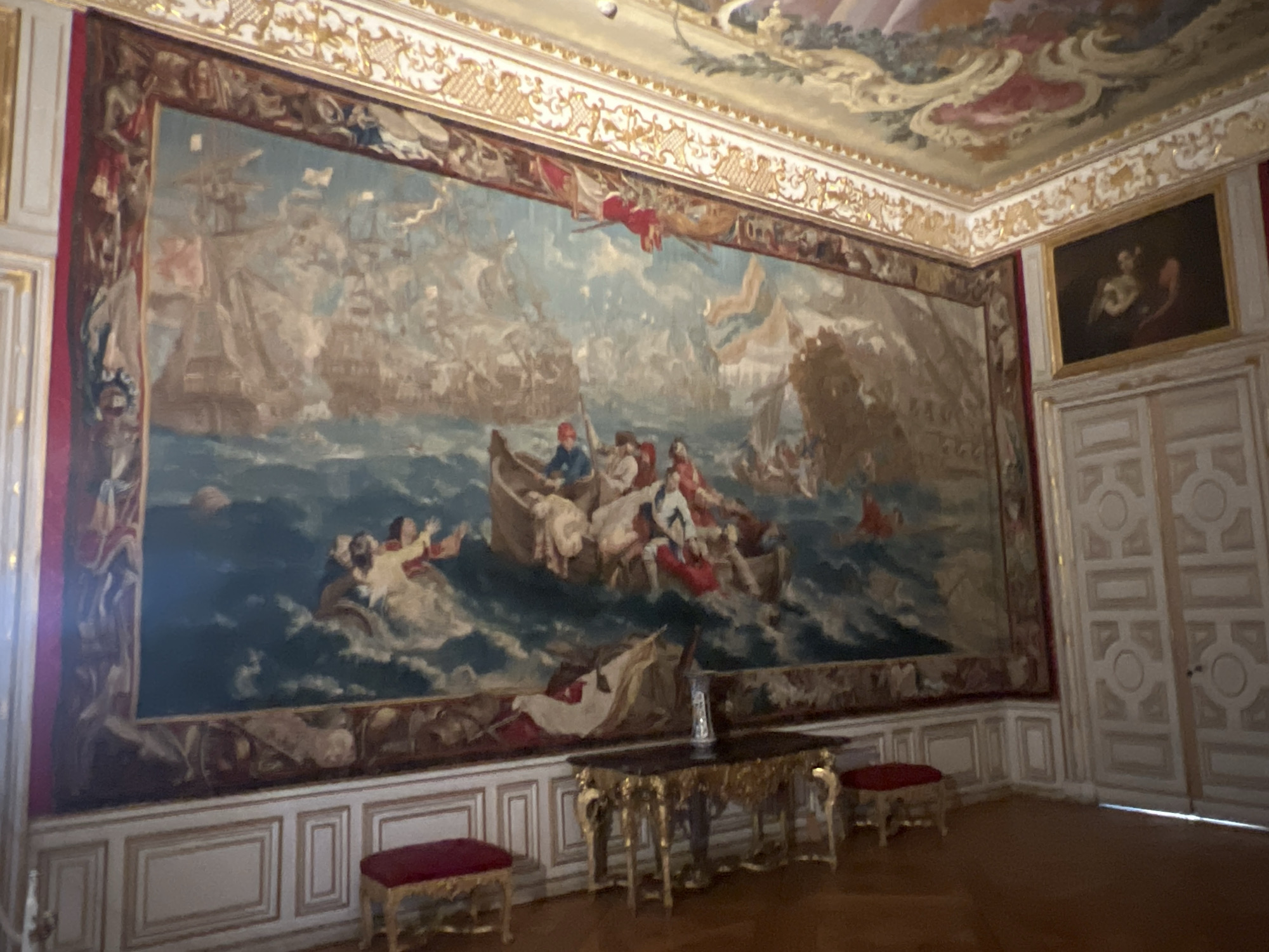 This is an indoor photograph of a grand hall in the New Schleissheim Palace, located in Oberschleißheim, Germany. The hall is dominated by a large, intricately woven tapestry depicting a naval battle scene with numerous ships and figures in boats amidst turbulent waters. The tapestry is framed by ornate gold trim and red fabric borders. Above the tapestry, the ceiling is adorned with elaborate frescoes and gilded stucco work. To the right of the tapestry, there is a framed oil painting of two figures on a dark background. The room features classic architectural elements, including paneled walls with gold detailing and a decorative frieze along the top. In the foreground, a golden ornate table with a dark top sits with a glass vase on it. Two small, rectangular stools with red velvet upholstery are placed in the room, one to the left and one to the right of the table. A large set of double doors with a decorative design is visible on the far right of the image. The floor appears to be polished wood.