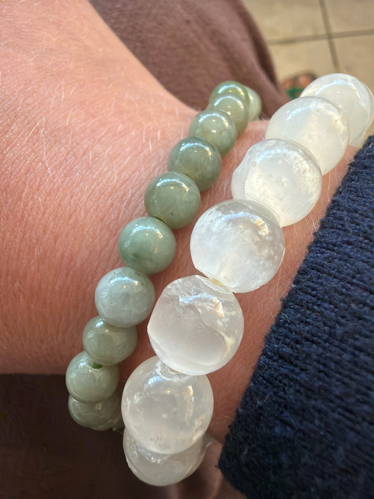 Two bracelets are displayed on a person's wrist. One bracelet features smooth, round, pale green beads. The other bracelet is composed of larger, irregular, translucent white beads. The person's skin, with visible pores and fine hairs, is partially visible. The background consists of a blurred brown garment and a dark blue knitted fabric. The provided image does not contain any information that would allow for the identification of its location in Pasay, Philippines.