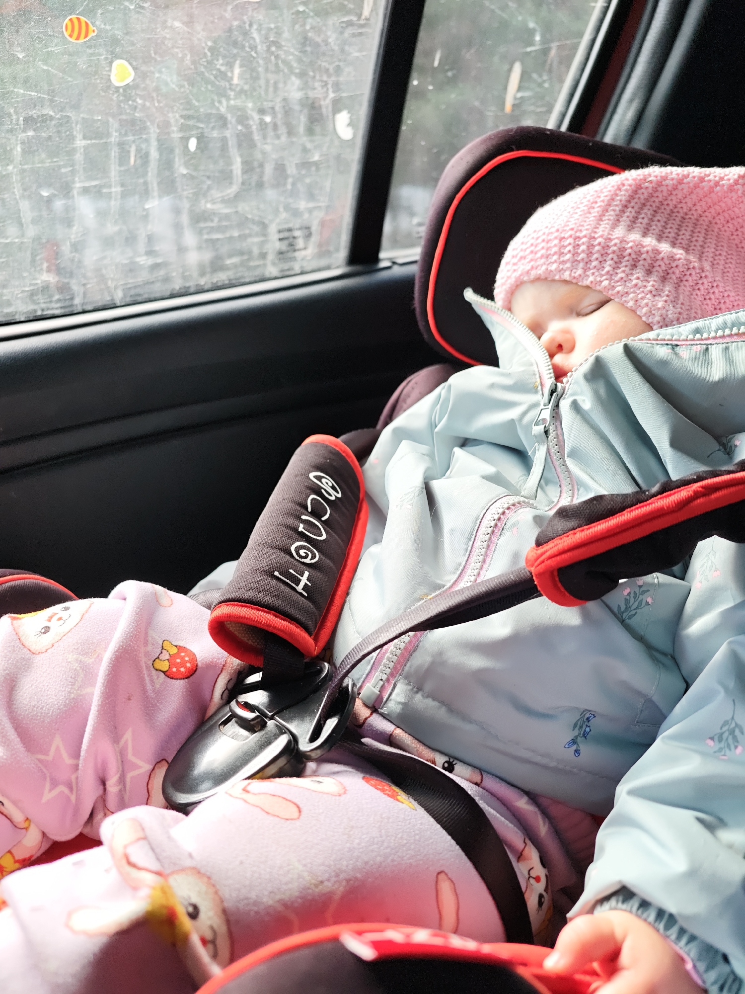 A baby wearing a pink knitted hat and a light blue jacket is sleeping in a car seat. The baby is secured with a harness. The car window is to the left, showing some indistinct shapes and stickers. One strap cover for the car seat has the word "SCOOT" written on it. The baby is also wearing pink pajamas with bunny prints. The scene appears to be inside a car, with the window showing a blurred outdoor view. There is no information available to determine the precise location as Chaikovsky, Russia from this image.