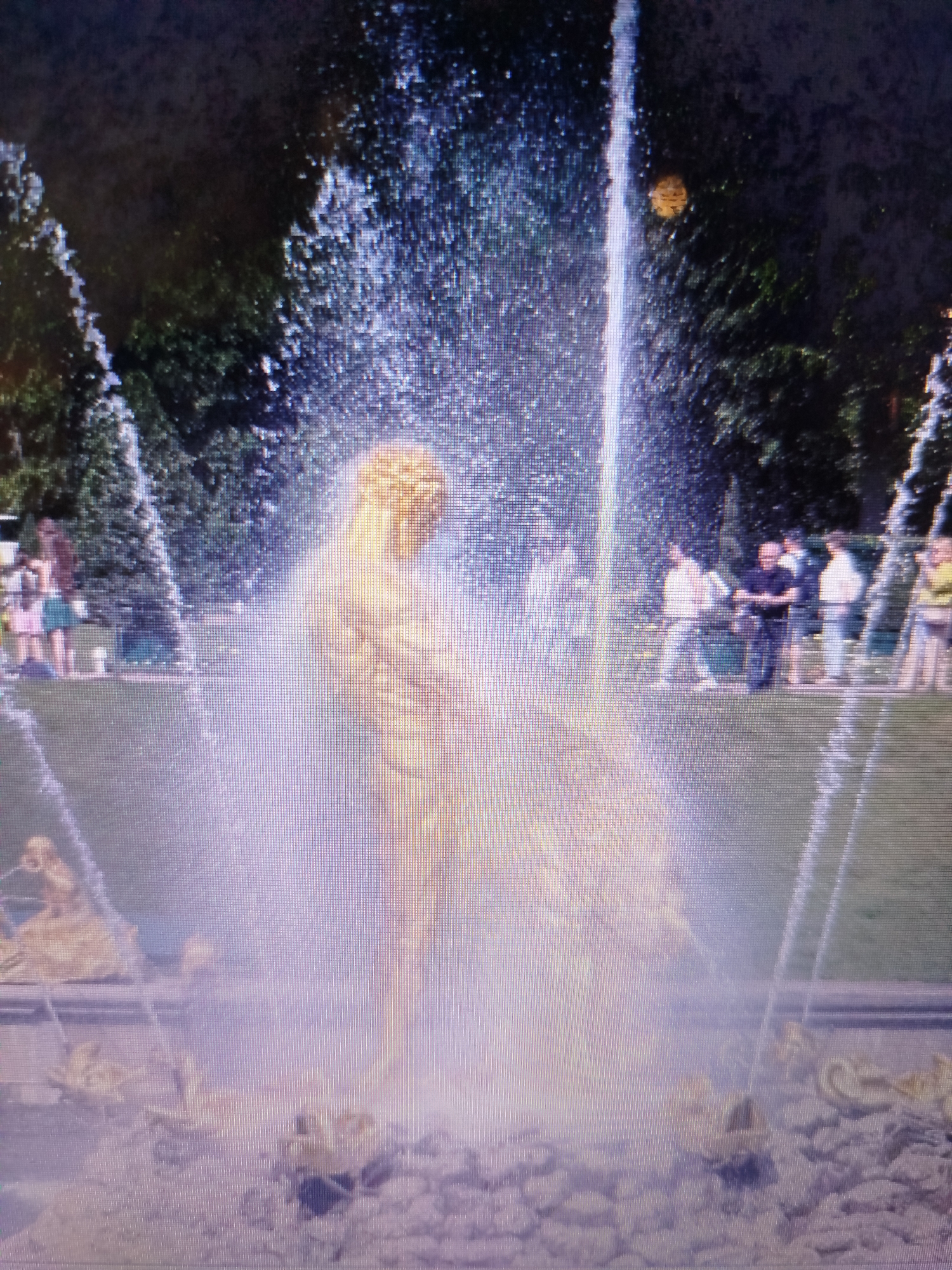 The image shows a golden statue of a muscular male figure, likely a mythological god or hero, as part of a large fountain. The statue is positioned centrally and appears to be the main subject of the fountain's elaborate water display. Water streams upwards from behind the statue and cascades down around it, creating a misty, ethereal effect. The statue itself is illuminated and appears golden, with intricate musculature visible despite the water's obscuring spray.
In the background, a park-like setting is visible with lush green trees and manicured grass. Several people are scattered throughout the background, standing and observing the fountain. They appear to be casually dressed, suggesting a public park or tourist attraction. Some individuals are standing in groups, while others are more isolated. The overall atmosphere suggests a sunny day, with the water from the fountain catching the light.
The statue is surrounded by a stone basin with more ornate sculptural elements at the base, also appearing to be in a golden finish, possibly depicting mythological creatures or decorative motifs. The ground around the fountain is covered with small stones. The image quality is somewhat grainy and appears to be a photograph taken at a distance, possibly with a phone camera.
The location is identified as the city of Snizhne, Ukraine. This suggests the fountain is a landmark or a feature within a public space in that city. The presence of trees, grass, and a fountain indicates a park or a garden area.