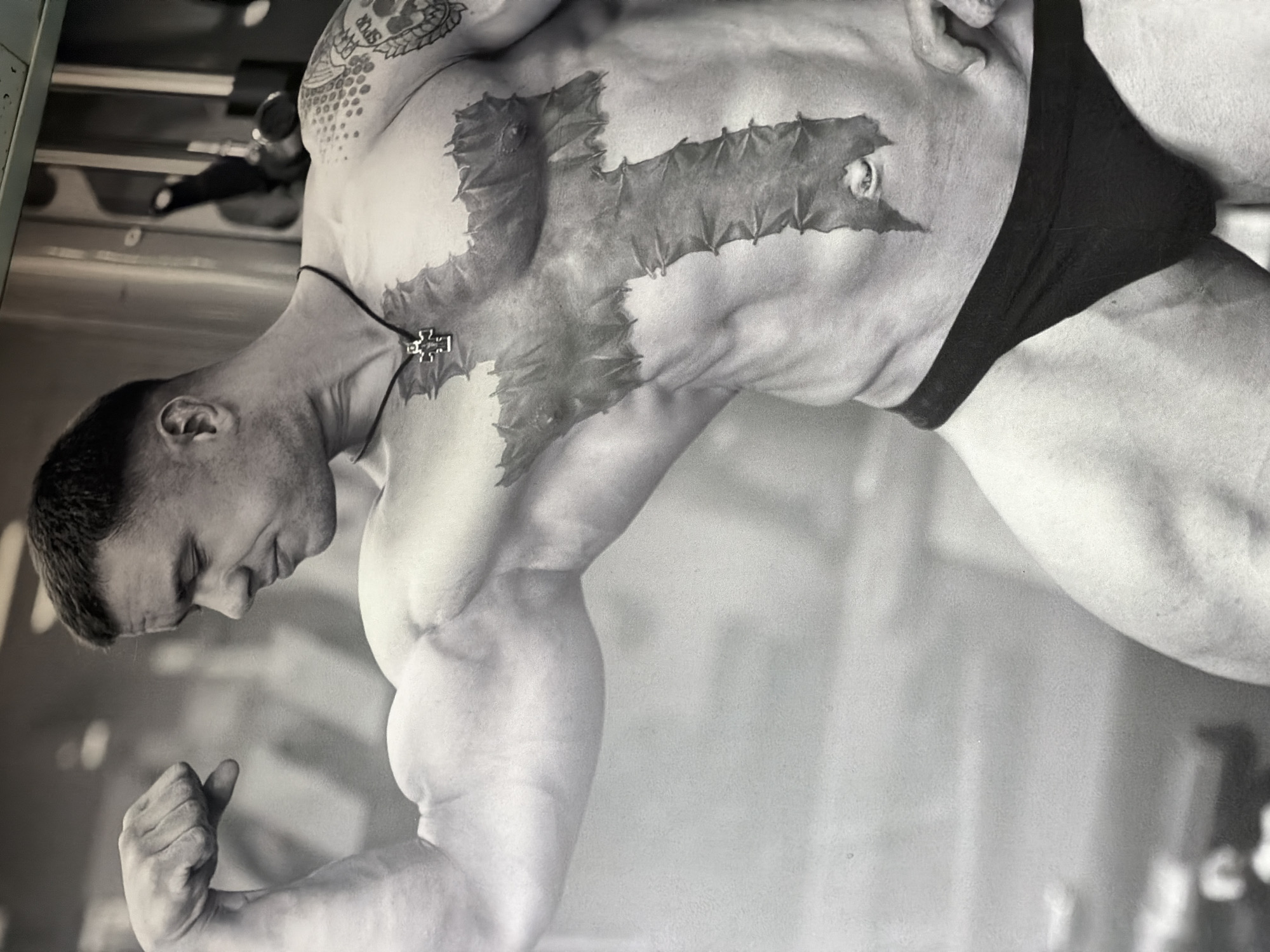 A black and white photograph depicts a muscular man, likely a bodybuilder, flexing his arm. He has a large cross-shaped tattoo on his chest and a smaller tattoo on his upper arm. He wears a minimal garment that appears to be underwear. The background suggests he is in a gym or fitness facility, with equipment partially visible. There is no information from the image indicating this photo was taken in Izhevsk, Russia.
