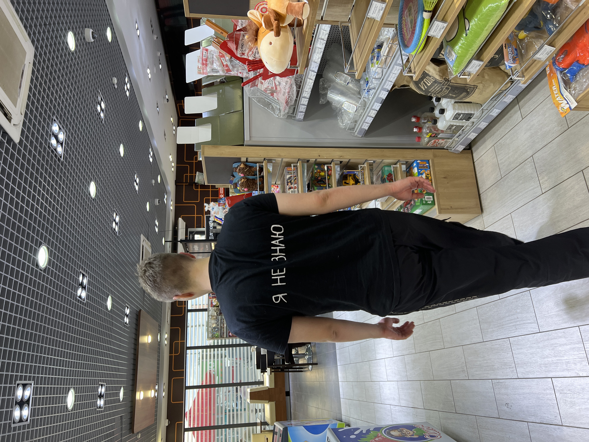 A man walks away from the camera down an aisle in a brightly lit convenience store. He is wearing a black t-shirt with white Cyrillic lettering that reads "Я НЕ ЗНАЮ" (Ya ne znayu), which translates to "I don't know." He is also wearing black pants. The store aisle is lined with shelves displaying various products, including drinks, toys, and household items. The flooring is made of light-colored tiles. In the background, through a window, cafe seating can be seen. The scene appears to be indoors, possibly a gas station convenience store, given the typical offerings visible on the shelves and the overall ambiance.