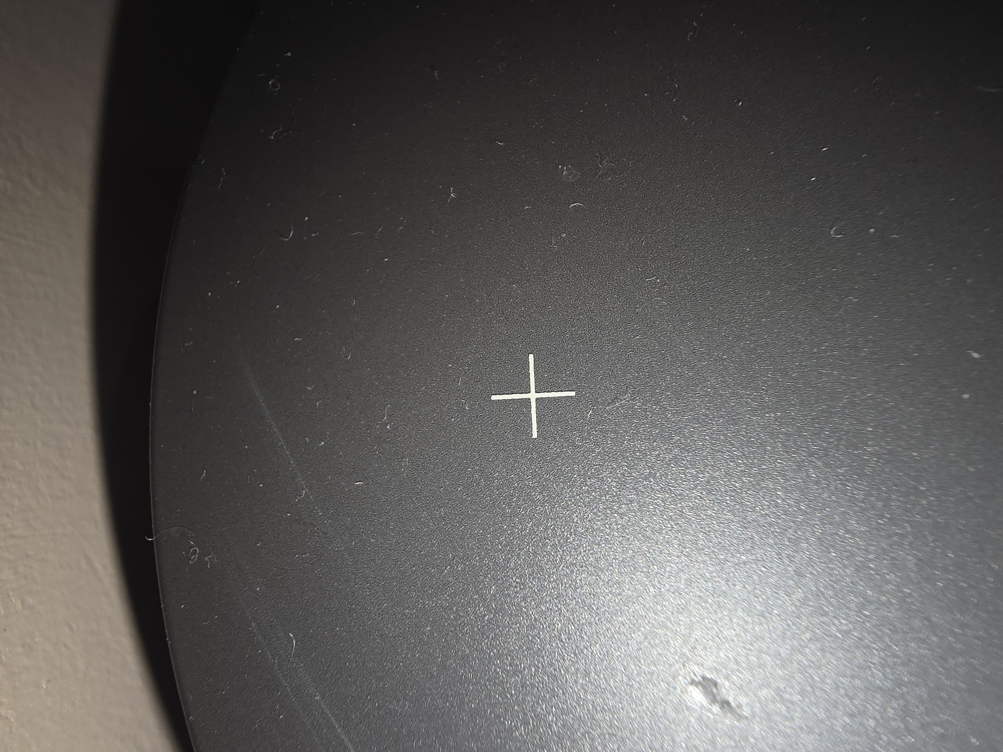 The image shows a close-up, angled view of a textured, dark gray surface with a thin white plus sign marked in the center. The surface appears to be a lid or outer casing of an electronic device, possibly a speaker or a piece of equipment. There are several minor scuffs, scratches, and small particles of dust or debris visible on the surface. The background is a blurred, light-colored wall with a shadow cast on it. There is no discernible information in the image that would allow for a determination of its location in Ulan-Ude, Russia.