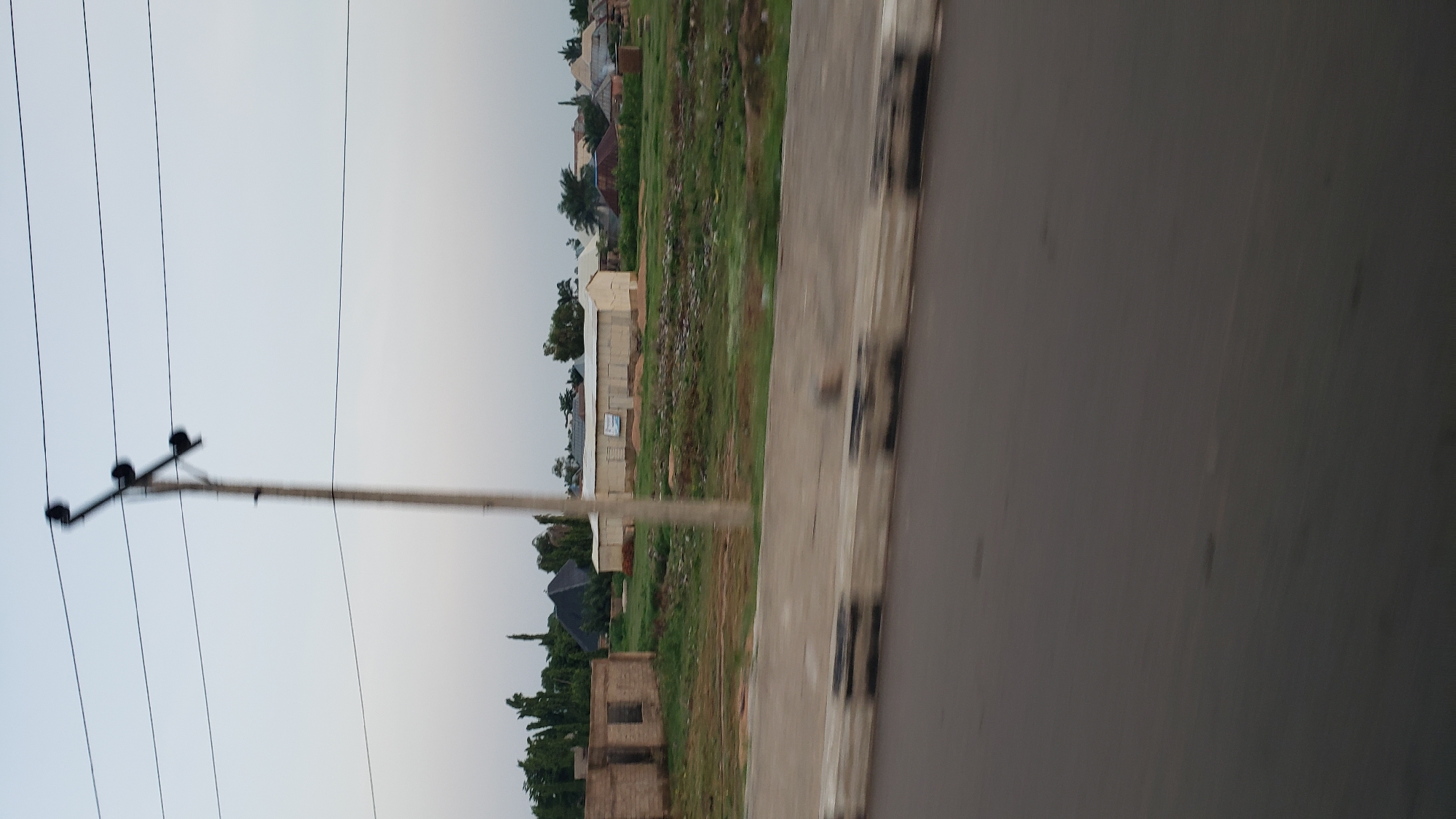 An eye-level, wide shot shows a street scene in Jalingo, Nigeria, during daylight. In the foreground, a paved road is visible, with a concrete barrier separating it from a grassy median. Beyond the median, a collection of buildings is present, including a partially constructed brick structure, a long, shed-like building with a white facade, and several smaller houses with corrugated metal roofs. A utility pole with power lines stands prominently in the middle ground, and lush green trees dot the landscape. The sky is overcast, with a pale, hazy quality. The image has a slight motion blur, suggesting it was taken from a moving vehicle.