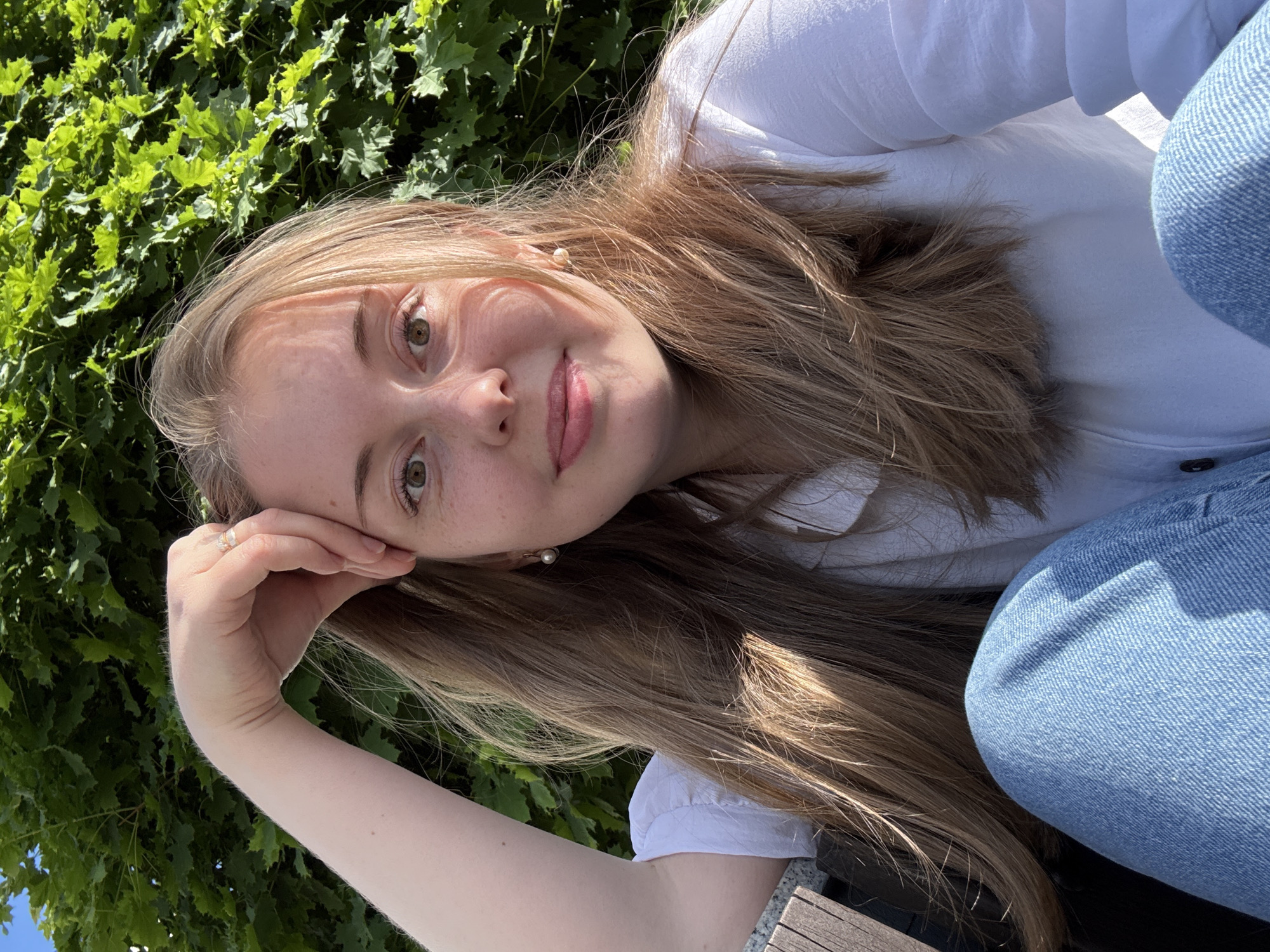 A young Caucasian female is depicted in a close-up outdoor portrait, seated and looking directly at the camera with a subtle smile. She has long, light brown hair with blonde highlights, brown eyes, and numerous freckles across her nose and cheeks. Her left arm is raised, with her hand resting lightly on her forehead; a multi-band gold-toned ring is visible on her left index finger. She wears a white collared button-down shirt and light blue denim jeans. Small, white pearl drop earrings are visible in both ears.
The background consists primarily of dense, bright green foliage, likely from a tree or large shrub, illuminated by strong sunlight, which also casts highlights and shadows across her face and hair. A sliver of clear blue sky is visible in the upper left corner. To the far left of the frame, a segment of a wooden structure, possibly a bench, is visible. The scene appears to be an outdoor public space in the city of Unknown, Unknown.