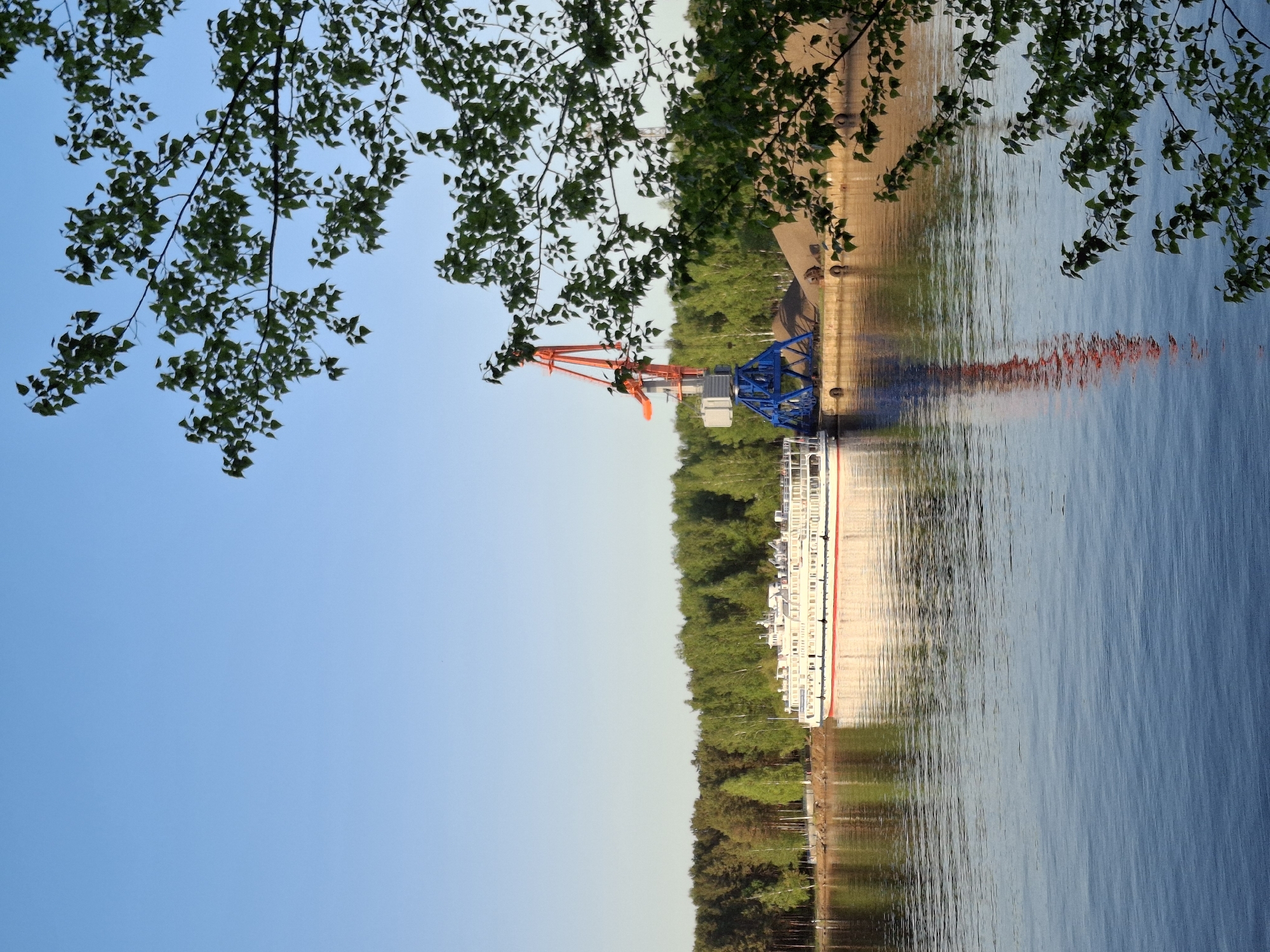 The image shows a view of a river in Chaikovsky, Russia. On the river, there is a docked boat with a large crane. The crane is painted red and blue. There are trees on the opposite bank of the river and along the shoreline. Branches with green leaves are visible in the foreground, framing the scene. The sky is blue with no clouds. The image seems to be taken with the camera rotated 90 degrees.