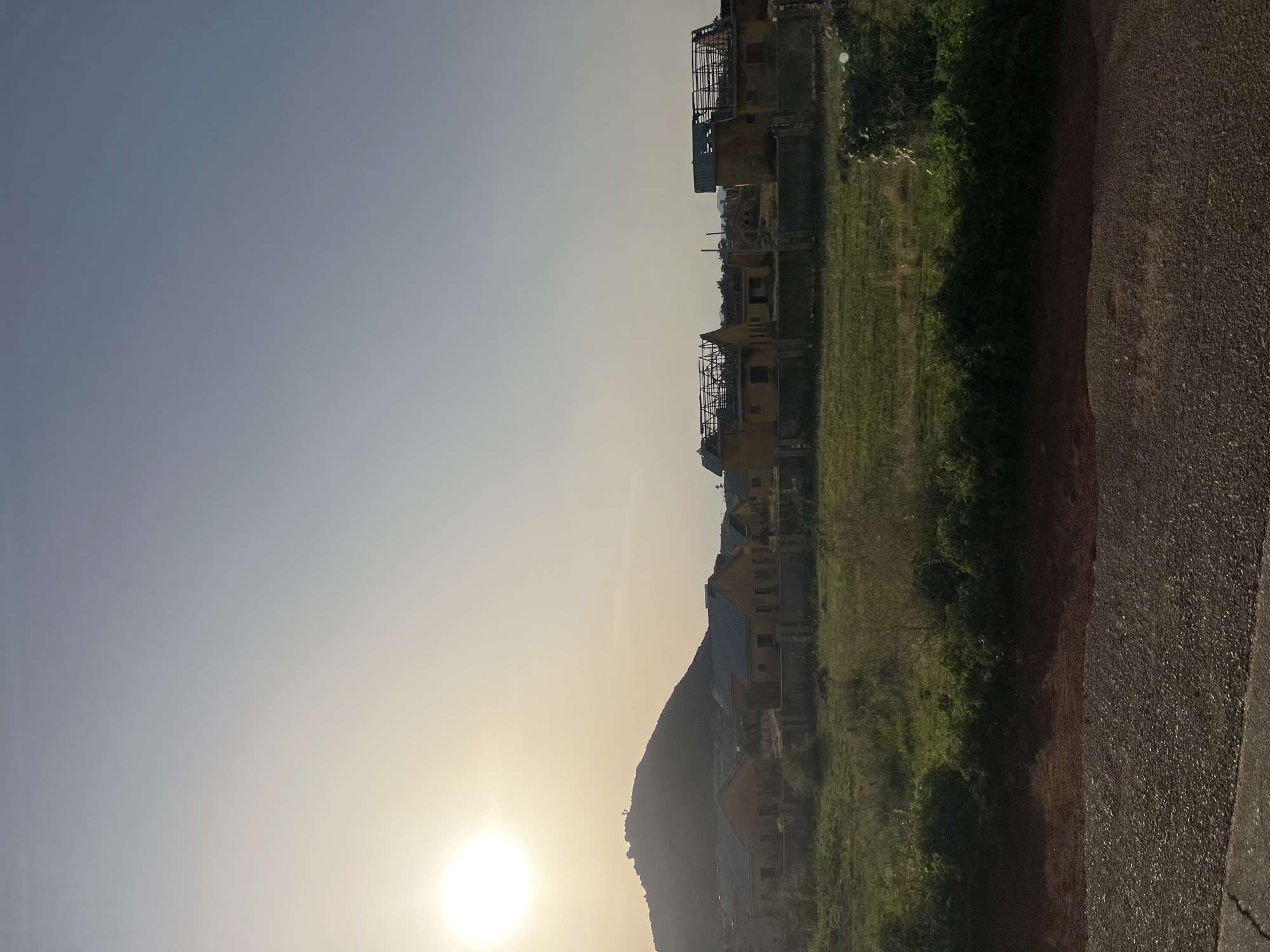 The image captures a wide outdoor scene in Bauchi, Nigeria, under natural light from a low-lying sun in the upper left quadrant, indicating either sunrise or sunset. A large, dark-silhouetted mountain or hill occupies the left-center background, partially obscuring the sun. In the mid-ground, a linear arrangement of approximately ten single-story structures, characterized by yellow-tan colored walls, is visible. Several of these buildings feature exposed roof trusses, suggesting incomplete construction or states of disrepair, particularly towards the right side of the frame. A low, grey concrete barrier or fence runs horizontally across the mid-ground, separating the structures from a foreground section of dense green grass and shrubbery. The immediate foreground consists of a dark, granular surface consistent with an asphalt or gravel road. No individuals or specific actions are discernible within the frame.