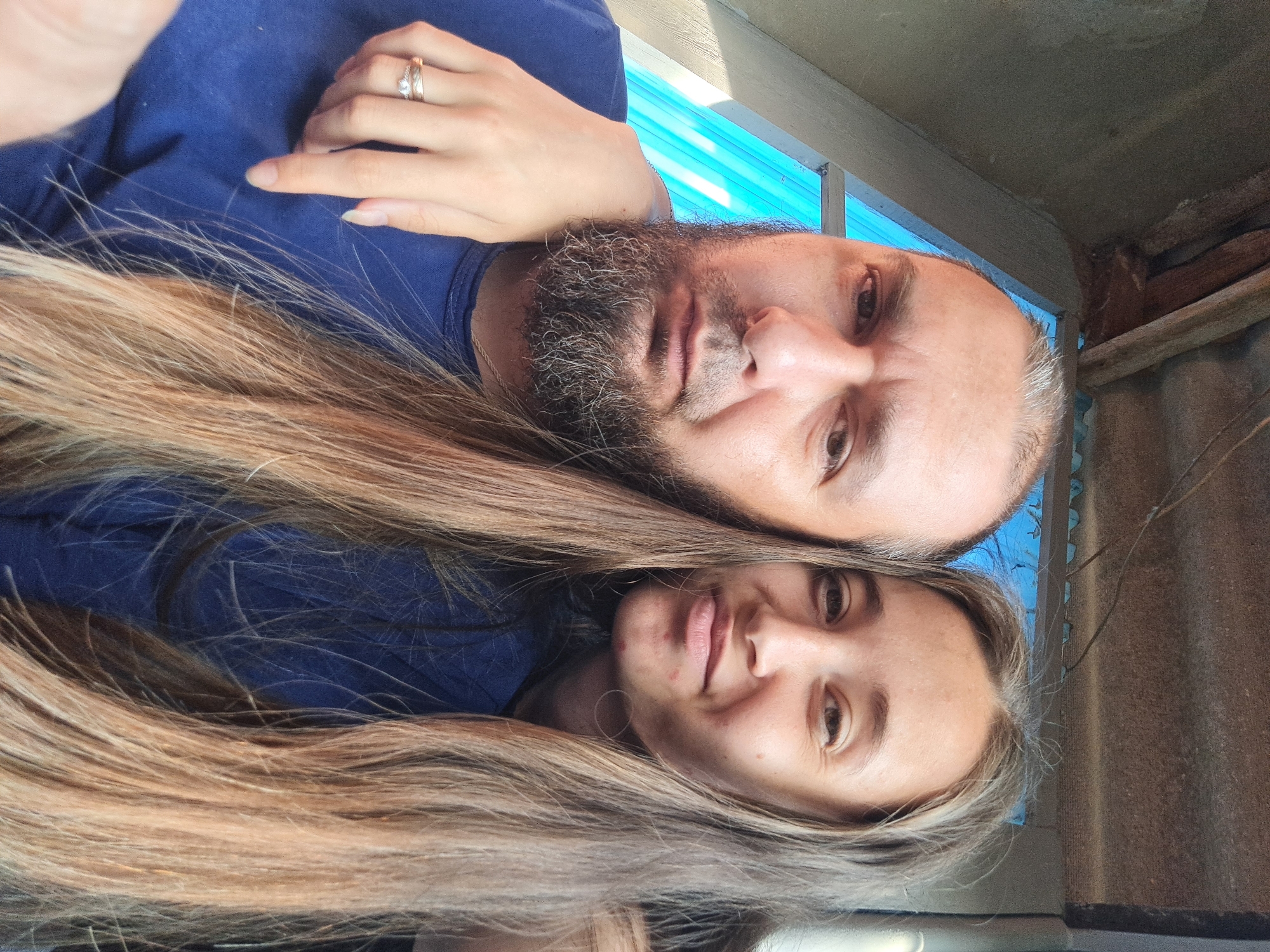 A selfie taken outdoors, possibly in Vypasne, Ukraine, features two individuals. The person on the left, a man with a full beard and long hair, is looking directly at the camera with a neutral expression. His right arm is around the shoulders of the person on the right, a woman with long, light brown hair. She is smiling slightly and her left hand, wearing a ring, rests on the man's shoulder. They are both wearing blue tops. In the background, a corrugated metal roof structure and a blue translucent material are visible. The overall lighting suggests it is daytime.