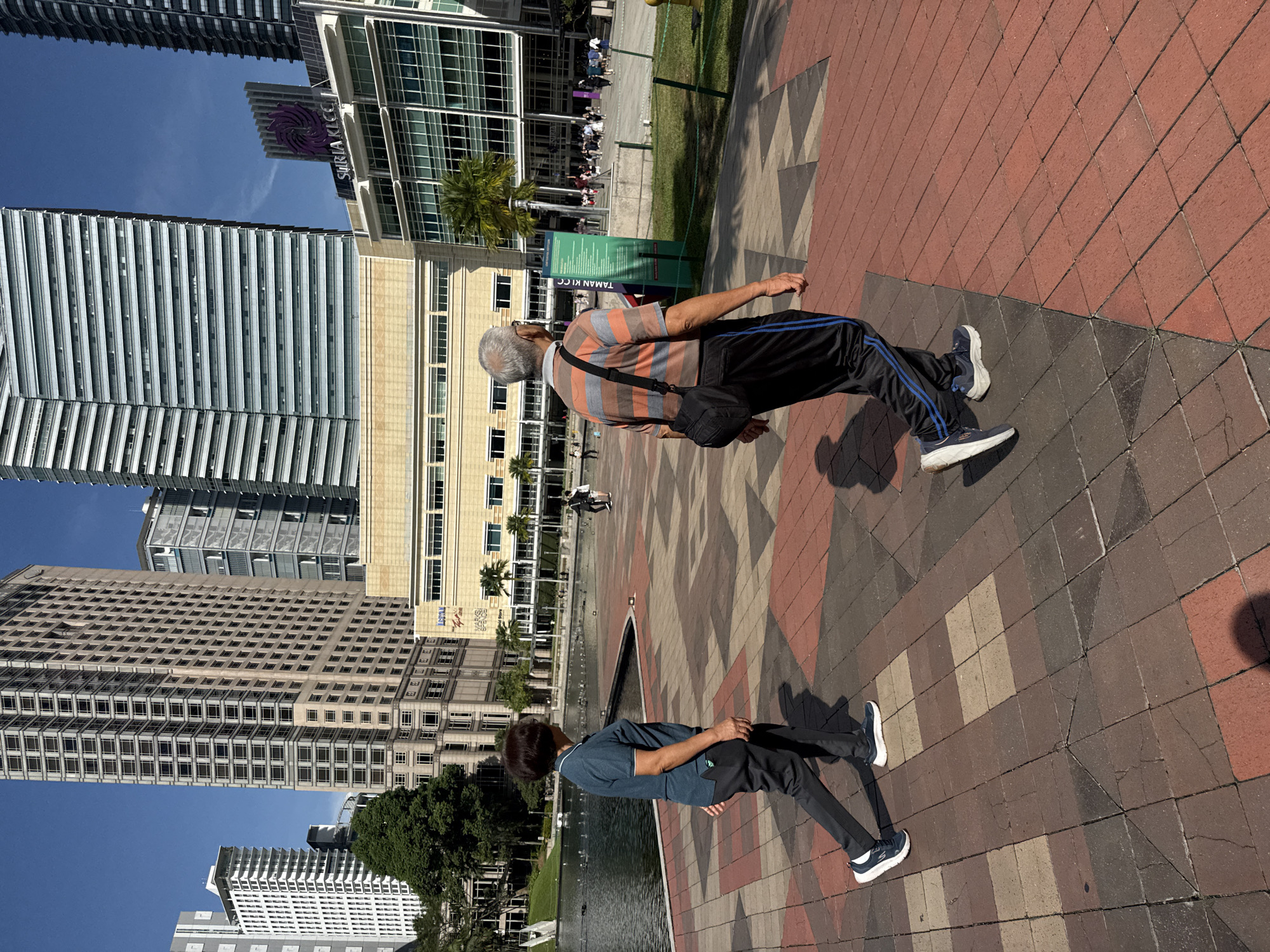 An outdoor scene depicts two men standing in Kuala Lumpur, Malaysia. The primary subject is an older man wearing a striped orange and grey shirt, black pants with blue stripes, and athletic shoes. He also has a crossbody bag. Adjacent to him, his reflection is visible in a body of water. He is also wearing athletic shoes, dark pants, and a blue shirt. The ground is covered with patterned red and tan brickwork. The background includes several high-rise buildings with modern architecture under a blue sky. The Suria KLCC shopping mall is visible. The environment appears to be a well-maintained public space with landscaping.