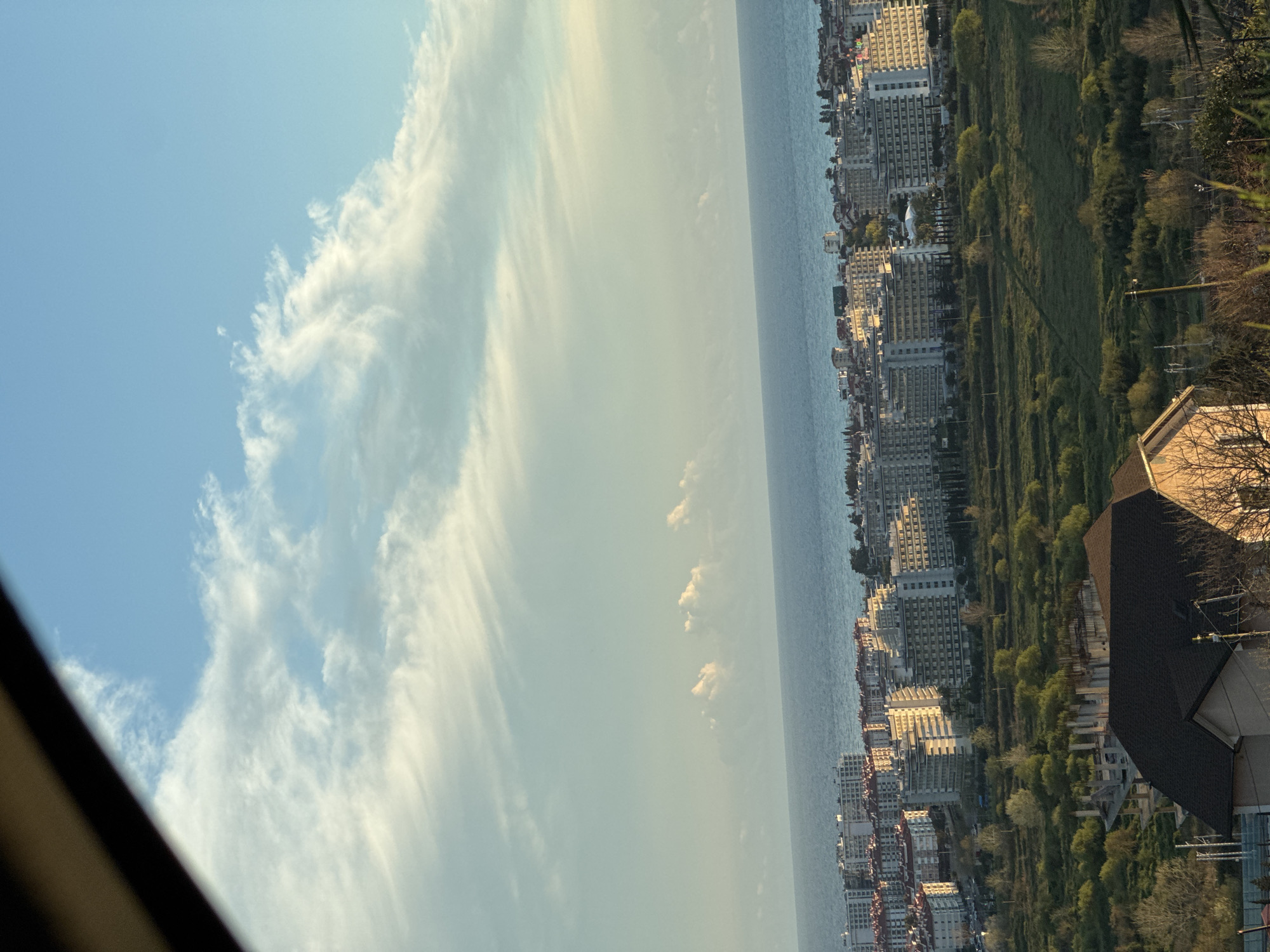 The image captures a view of Sochi, Russia, displaying the city's coastal landscape. On the right, dense clusters of buildings, including high-rise structures, are visible, indicative of urban development. Adjacent to the buildings, green spaces with scattered trees suggest parks or landscaped areas. The sea stretches horizontally across the middle ground, and the sky occupies the remaining space. The sky is partly cloudy with varying cloud formations, from scattered puffy clouds to more diffuse, streaky cloud patterns. The color palette is dominated by blues and greens, complemented by the muted colors of the buildings and vegetation.