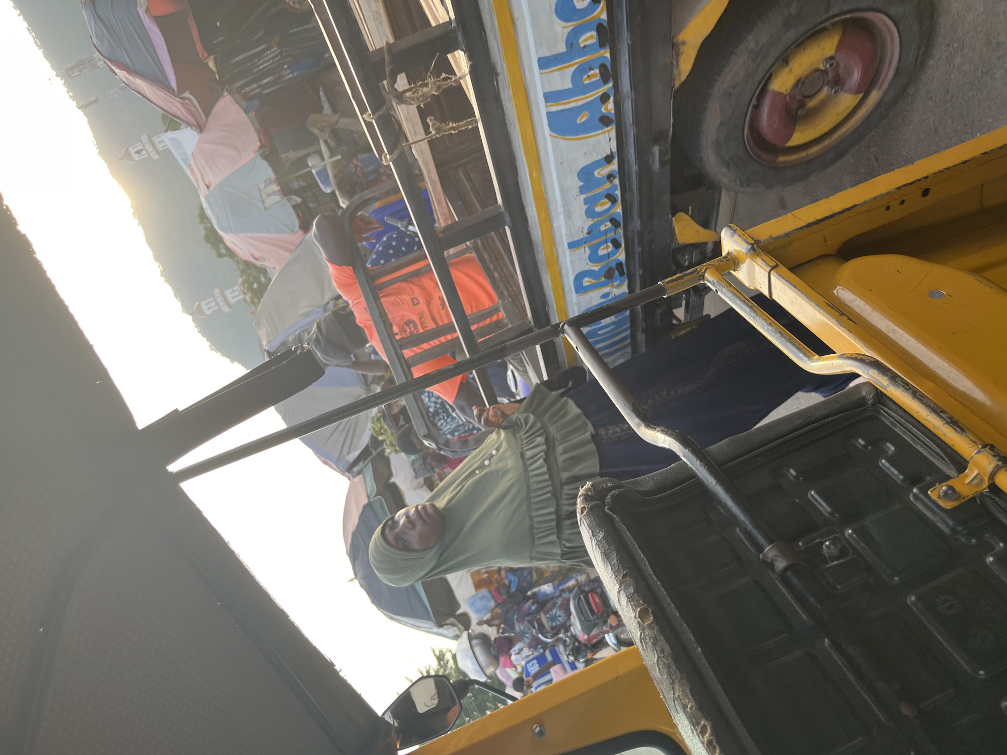 In the city of Unknown, Unknown, a woman wearing a green hijab and a dark blue skirt stands beside a yellow auto rickshaw. She is looking to her right, with her right hand slightly raised. Behind her, a man in an orange shirt leans against the railing of a truck. The truck has "Baban Abba" written on its side in blue and yellow letters. In the background, market stalls with colorful umbrellas are visible, along with a distant mountain and two minarets. The sun is setting, casting a warm glow over the scene.