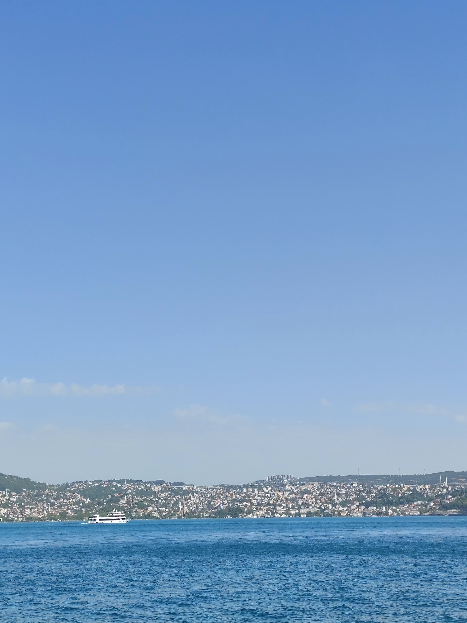 The image shows a distant view of Sarıyer, Turkey, from across the Bosphorus Strait. The scene consists of blue water in the foreground, with the opposite shoreline densely populated with buildings and greenery. The sky is a clear, light blue with a few scattered clouds. A white ferry is visible on the water near the shore. In the background, the cityscape is a mix of residential and commercial buildings scattered across the hilly terrain.