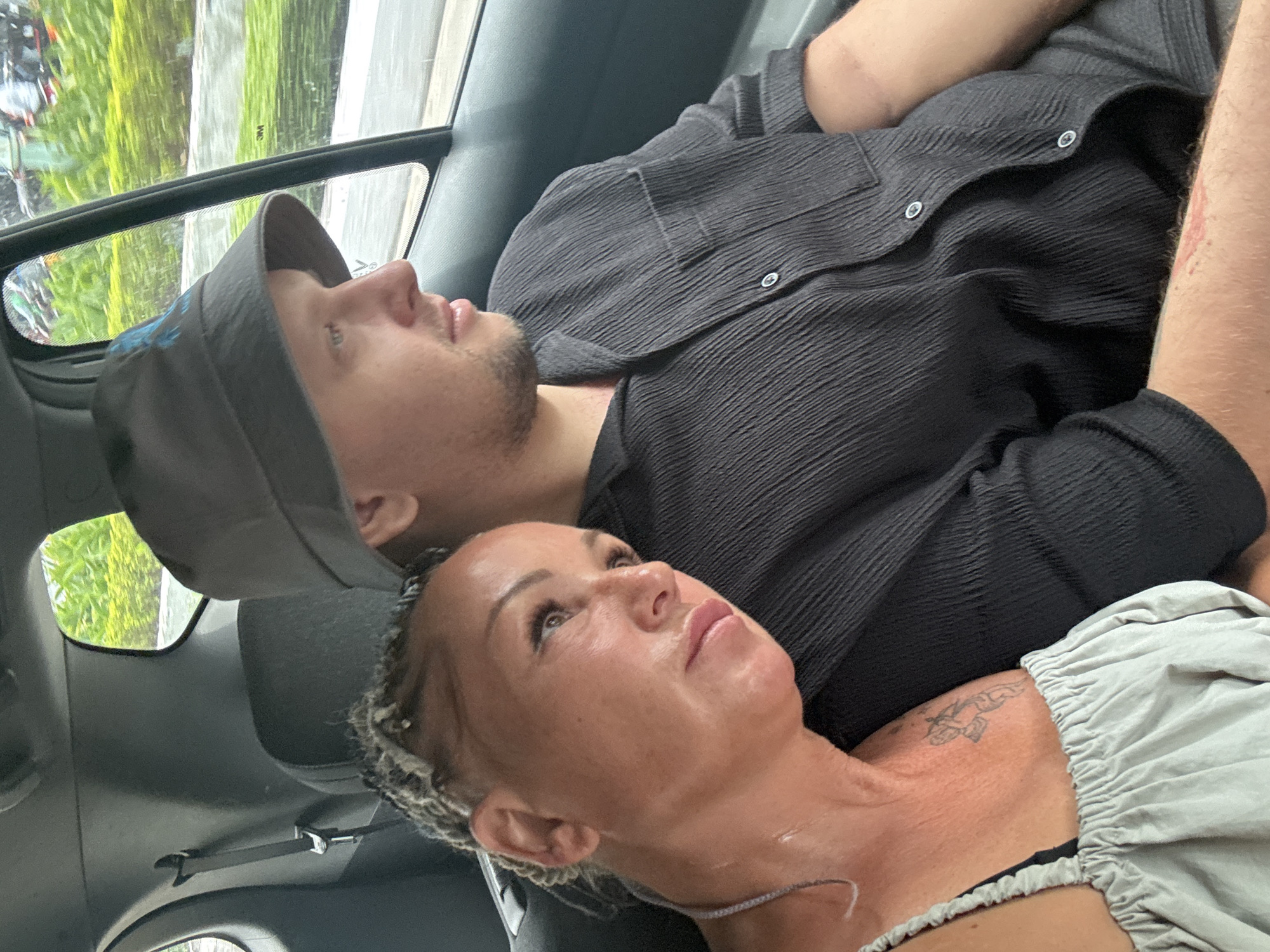 Inside a vehicle, a man and woman are visible. The man wears a dark gray hat and a black textured shirt. He has a beard and is lying back. The woman is in the foreground, also lying back, with her head tilted upwards. She has braided hair and a tattoo on her upper chest/neck area. The interior of the vehicle is dark. A window shows an out-of-focus green landscape, suggesting movement. The setting appears to be Đà Nẵng, Vietnam, based on the vegetation seen through the window.