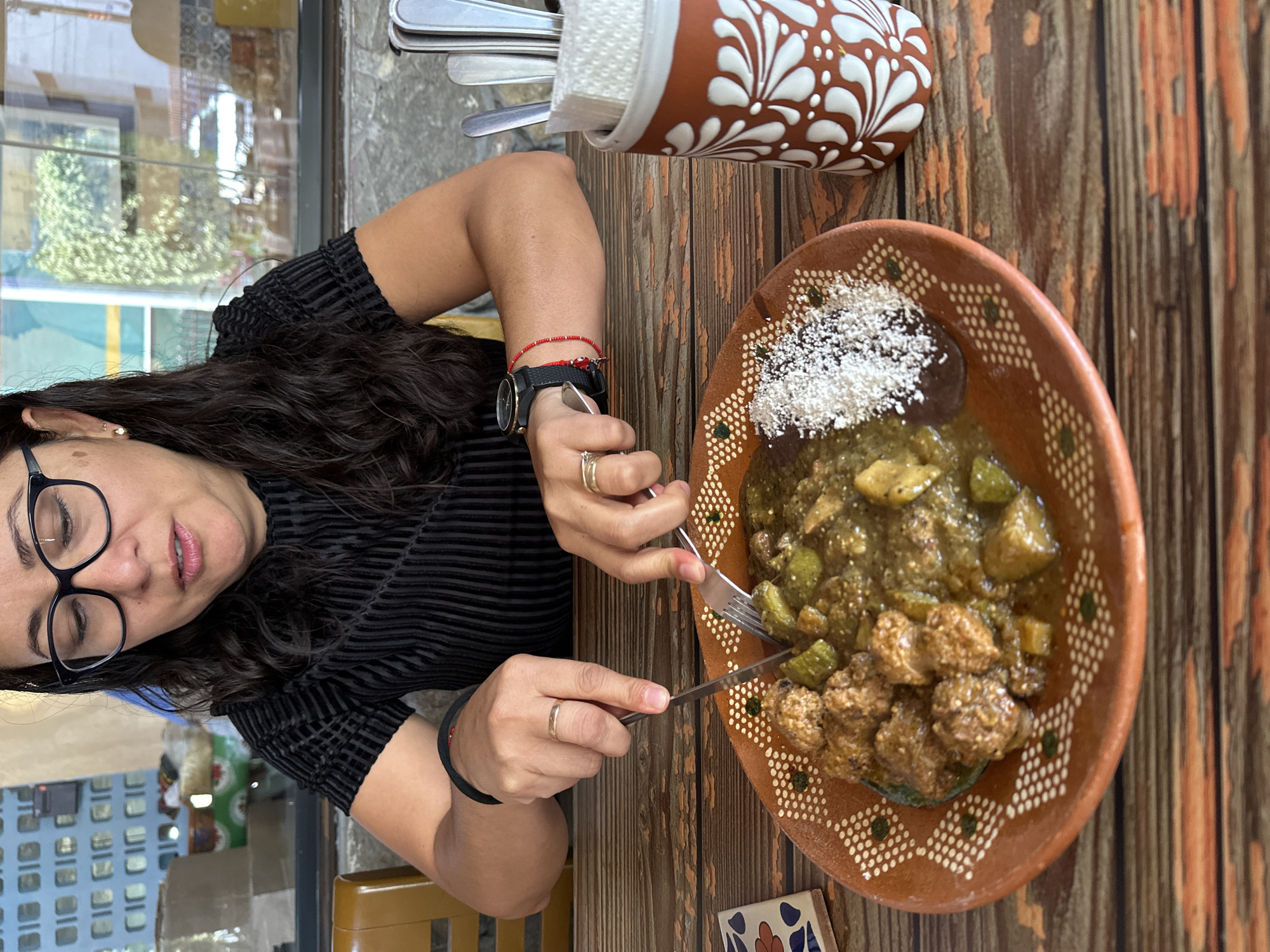 A woman with dark hair and glasses is sitting at a wooden table in what appears to be a restaurant, likely in Puebla, Mexico, as indicated by the traditional Talavera-style plates and possibly the architecture visible in the background. She is wearing a black, textured shirt and a watch. Her left wrist has a red beaded bracelet. She is using a knife and fork to eat a meal from a brown Talavera plate. The plate contains what appears to be green salsa with chayote, accompanied by what might be meatballs and a dollop of beans topped with cheese. A Talavera container holding utensils sits to the top of the frame. The table is made of vertical wooden planks.