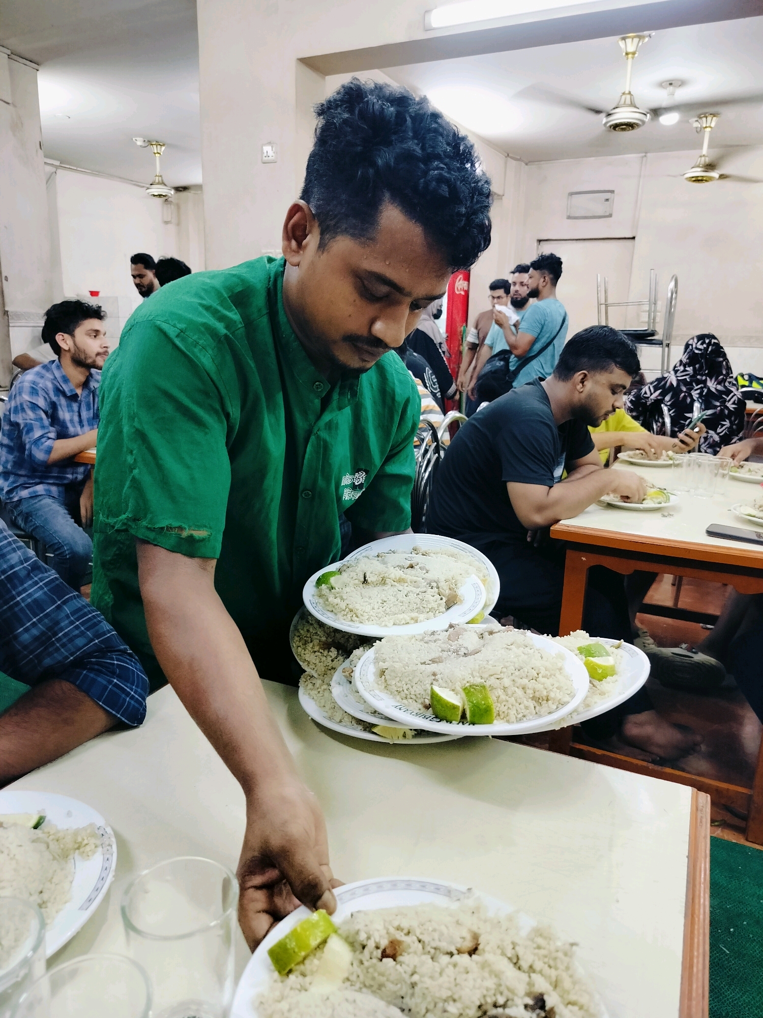 The image depicts a casual dining scene in a restaurant, likely in Dhaka, Bangladesh. A server, wearing a green shirt with visible tears on the sleeve, is in the foreground, holding plates of food. The food appears to be rice with some meat, garnished with lime wedges. Several customers are seated at tables, engaged in eating. One man in a blue plaid shirt is looking to his left, while others are focused on their meals or phones. The environment suggests a modestly furnished eatery with ceiling fans visible on the white ceiling. The overall impression is that of a busy, everyday mealtime at a local establishment.