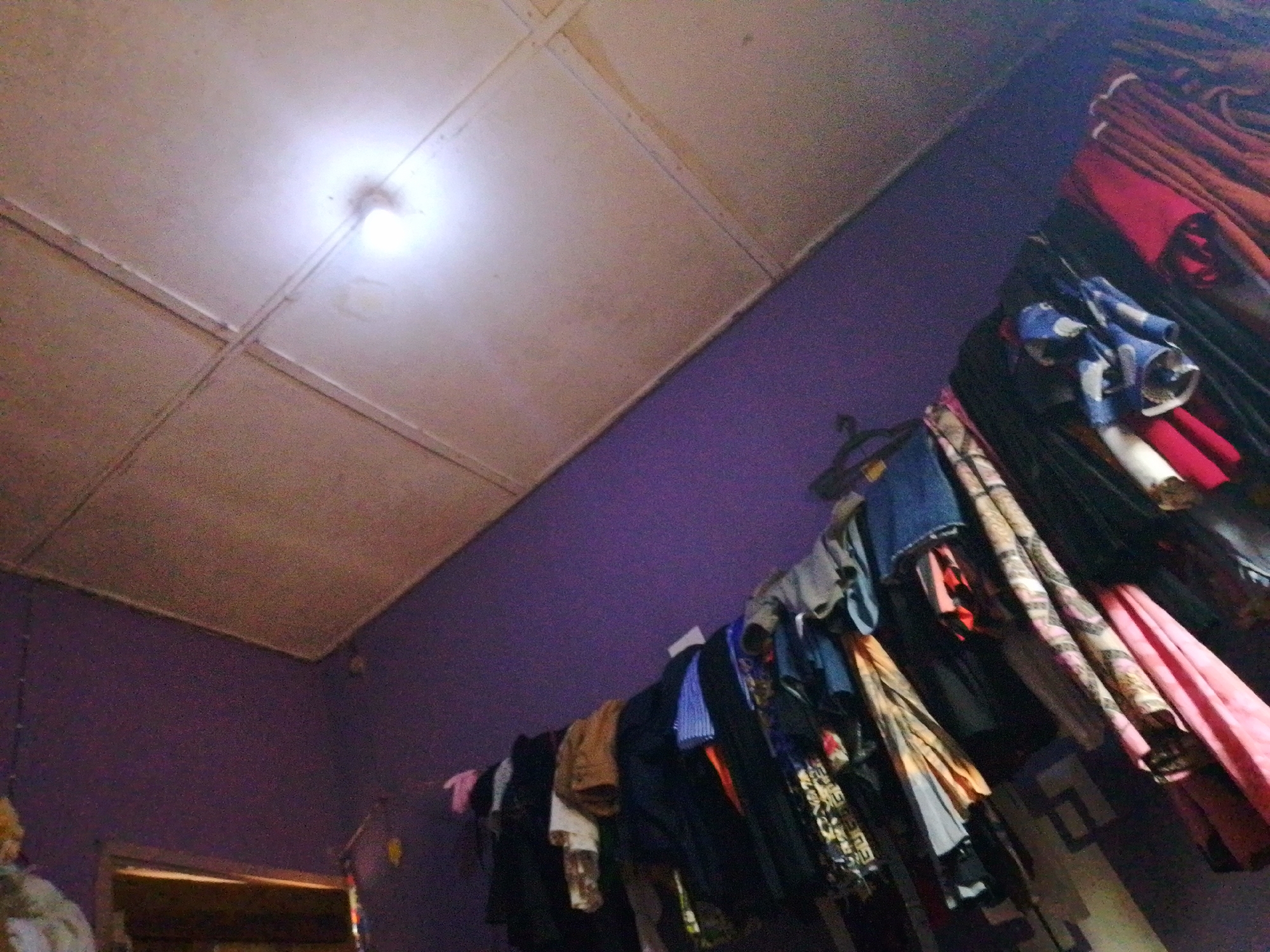The image depicts a room interior, possibly in Nsukka, Nigeria. The scene is illuminated by a single overhead lightbulb, which casts a bright glow on the textured ceiling panels. The walls are painted a deep purple. A clothesline, or similar hanging apparatus, is visible across the upper part of the room, laden with various items of clothing. These include what appear to be shirts, pants, and potentially some patterned fabrics. The items are hung haphazardly, with some folded and others draped. The lower portion of the image shows the wall continuing downwards, with a dark doorway or entrance visible on the left. The overall impression is of a functional, lived-in space, likely a bedroom or living area, where clothing is stored by hanging.