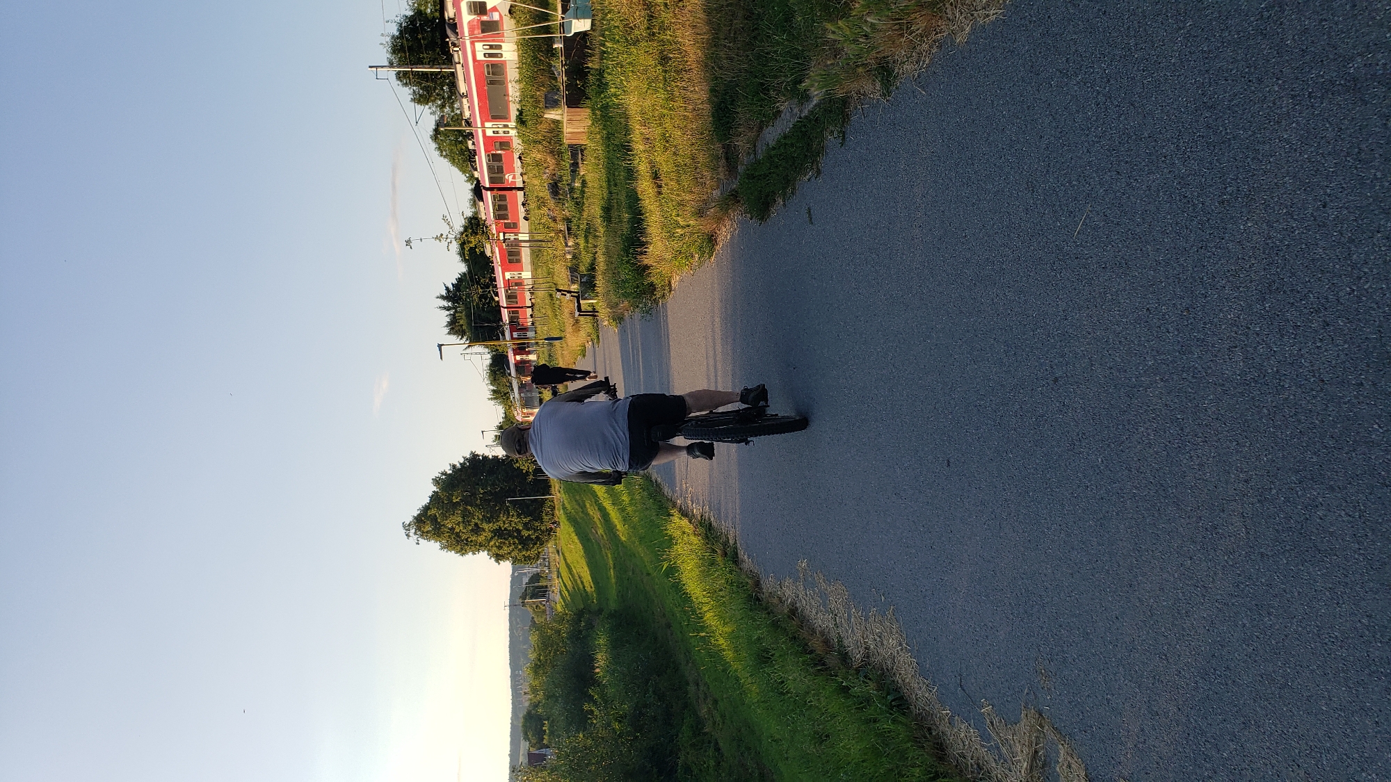 The image shows a person riding a bicycle on a paved path next to a grassy area. A red and white train is visible in the background, parked on tracks. The scene appears to be outdoors during the daytime, with a clear sky and sunlight casting shadows. The cyclist is wearing a grey t-shirt and black shorts, with a helmet on. They are riding away from the camera, with their back to the viewer. The path is dark grey asphalt, and the grass is green and lush. The train is long and modern, with multiple cars. There are some trees and vegetation on the left side of the image, and a hint of a town or city in the distance. The overall mood is peaceful and active, suggesting a leisurely bike ride on a pleasant day. The time of day appears to be late afternoon or early evening, given the angle of the sun. There is no visible text in the image.