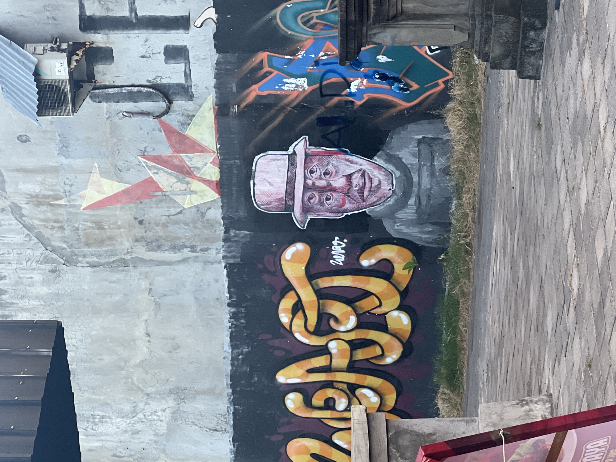 The image shows a wall with graffiti in Sukawati, Indonesia. The wall is divided into sections. The leftmost section is plain grey, with an air conditioning unit attached near the top and a geometric design in red, yellow, and white below. The center section of the wall is black and contains various graffiti elements, including a blue and orange design at the top, a drawing of a person's face wearing a hat, and the word "zuvot" written in yellow and orange with a white outline. To the right of the graffiti, there is a narrow strip of grass growing along the edge of the wall, where it meets a paved sidewalk. The edge of a building is visible in the foreground at the bottom of the image. The overall condition of the wall appears weathered and worn.