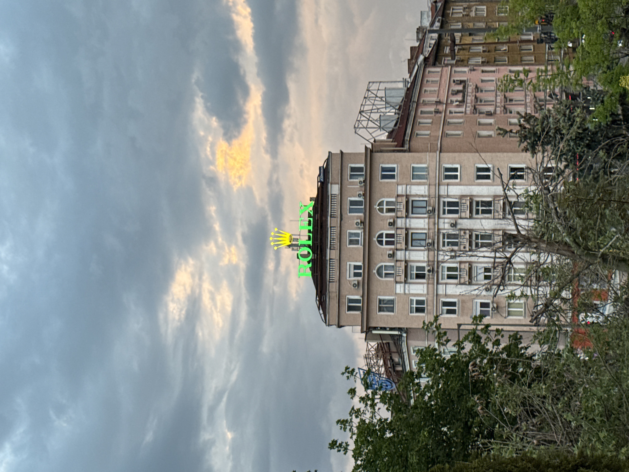 The image depicts a building with a Rolex sign on its roof, captured against a cloudy sky in Sofia, Bulgaria. The building is multi-storied, tan-colored, and features rows of windows. A green neon "ROLEX" sign and the Rolex crown are positioned on the building's top edge. Adjacent to the building is another structure, appearing older and with varied pastel hues. Trees with dense foliage frame the building on the right and in the foreground. The sky is filled with gray clouds, partially illuminated by a soft, yellowish light source, suggesting either sunrise or sunset.
