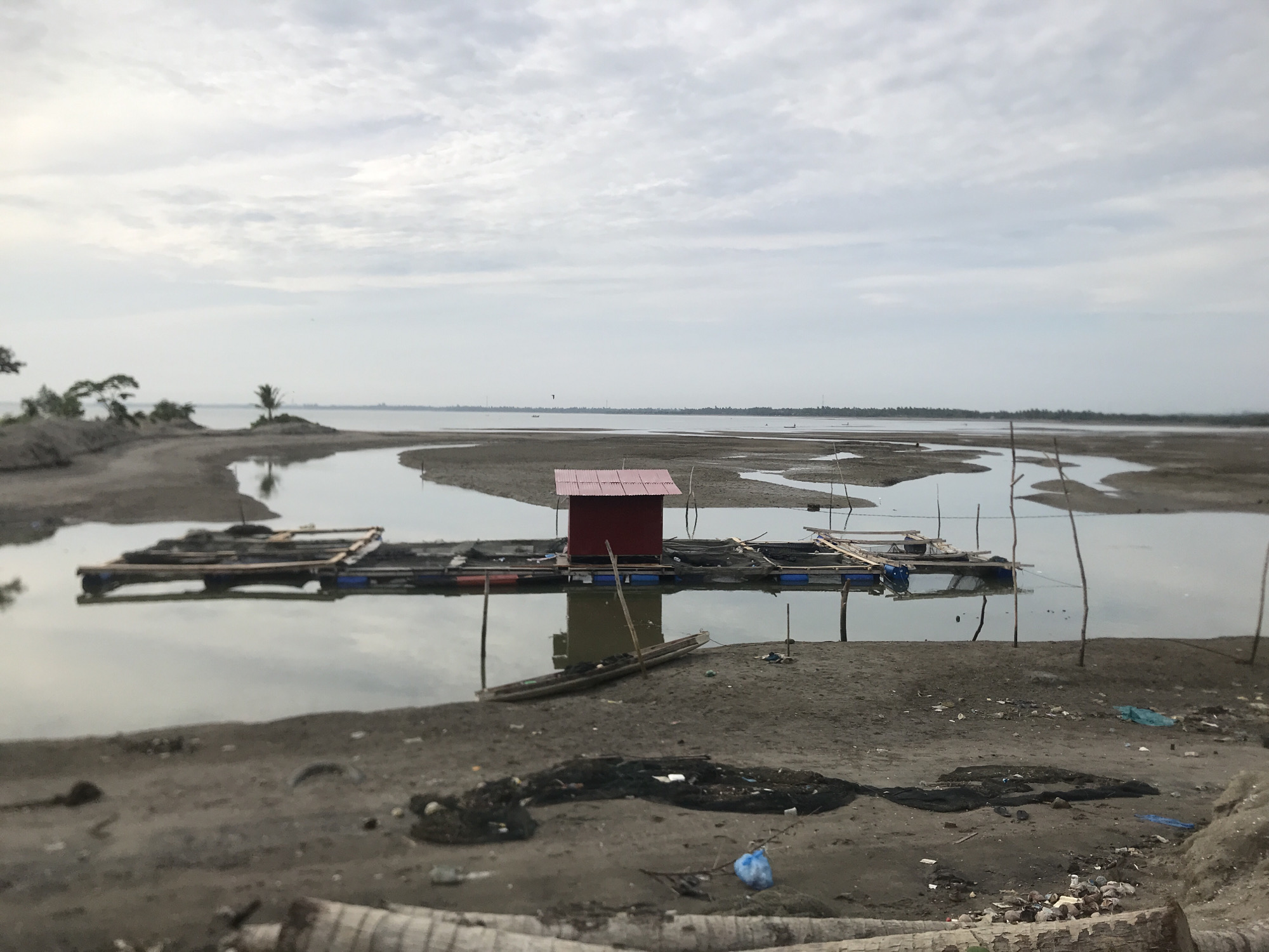 The image depicts a body of water with a muddy shoreline, likely a river or a coastal area in Lhokseumawe, Indonesia. A floating structure, possibly a fish farm or a small dwelling, is visible in the water. It consists of a red-roofed structure supported by wooden beams and plastic barrels. Several smaller wooden structures and a small, empty boat are also present in or near the water. The shoreline is covered with sand, debris, and some vegetation in the distance. The sky is overcast with gray clouds, suggesting a subdued or cloudy day. The overall scene conveys a sense of a rural or undeveloped waterfront, with signs of human activity related to fishing or aquaculture.