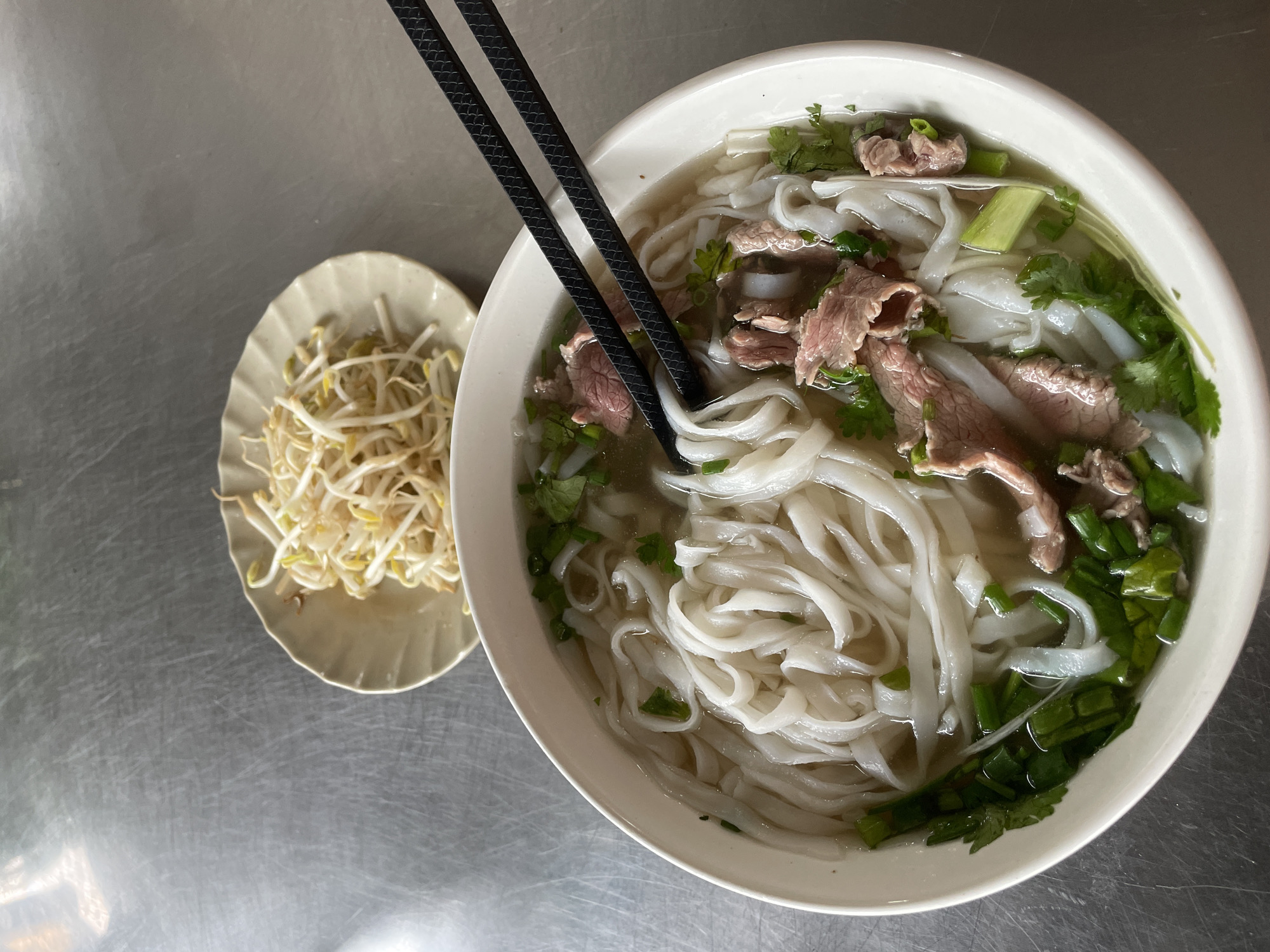 A bowl of pho, a traditional Vietnamese noodle soup, is displayed on a metallic surface. The pho contains rice noodles, thinly sliced beef, and green herbs such as cilantro and scallions. A pair of black chopsticks is positioned above the bowl, ready to pick up the noodles. To the upper right of the pho, a small dish of bean sprouts is placed on the same metallic surface. The scene appears to be set in a casual dining environment in Đà Nẵng, Vietnam.