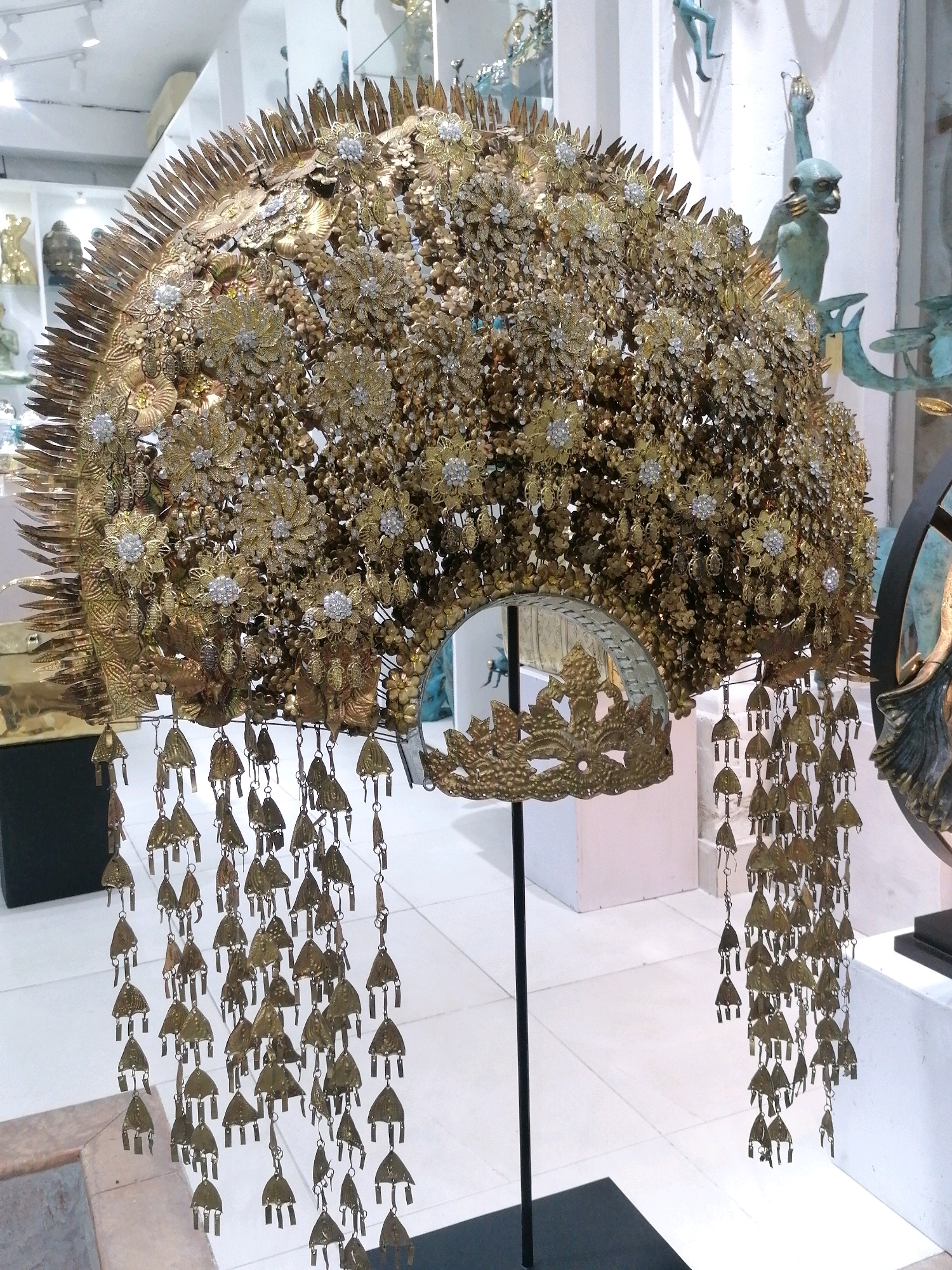 An ornate, large, golden headdress, possibly a ceremonial crown, is displayed indoors on a black stand. The headdress is intricately crafted with numerous small gold flowers, some adorned with clear rhinestones, and larger, more elaborate gold floral designs. Along the bottom edge of the headdress hang many strands of small, triangular gold ornaments. A curved, decorative gold piece with leaf-like cutouts is visible at the bottom of the headdress, partially obscuring a reflective surface, possibly a mirror. In the background, other decorative objects and sculptures are visible, including a turquoise statue of a monkey-like figure in a dynamic pose. The setting appears to be a gallery or museum, with white walls and flooring, and overhead lighting. The specific location in the city of Ubud District, Indonesia is not discernable from the image alone.