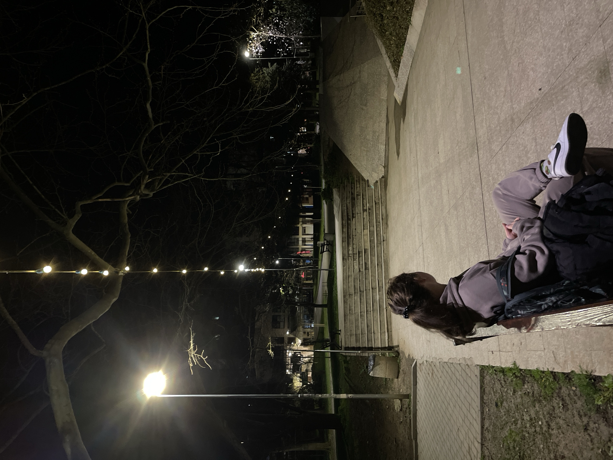 A woman is sitting on a bench at night in what appears to be a park or public space in Veliko Tarnovo, Bulgaria. She is wearing light-colored sweatpants and a sweatshirt and white sneakers with a black Nike swoosh logo. She has dark hair pulled back in a ponytail and a black backpack. The background includes bare trees, some with string lights. There are street lights in the distance, and concrete stairs are visible leading up a small incline. The environment suggests a quiet, urban outdoor setting.