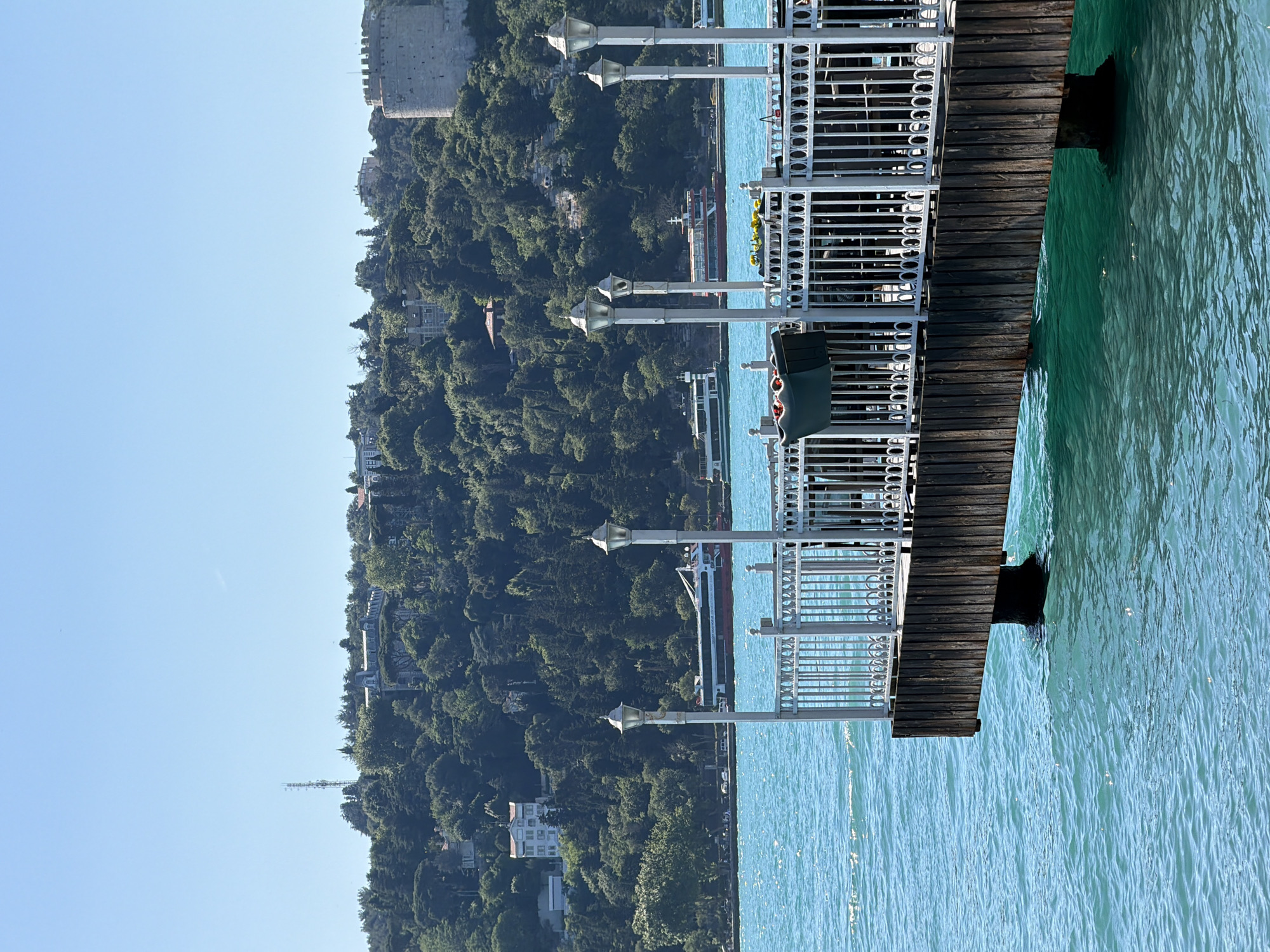 The image captures a view from a wooden pier extending into a body of turquoise water, likely the Bosphorus Strait, in Beykoz, Turkey. The pier has a white ornate metal railing and several lampposts. Beyond the pier, on the shore and rising hills, are numerous green trees. Several buildings are scattered amongst the foliage, and a prominent tower with battlements, likely a historical fortress, stands on the right side of the frame. In the distance, on the water, are two ferries or boats. The sky is a clear, bright blue. The scene appears tranquil and scenic, depicting a sunny day with calm water.