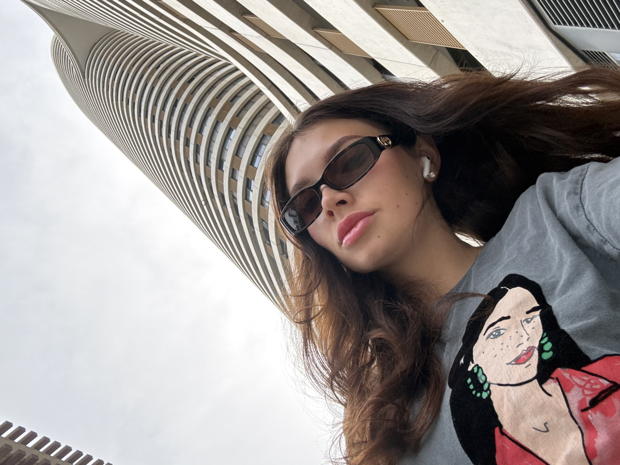 A young woman with long brown hair and sunglasses is pictured in a low-angle shot, with a tall, curved building in the background. She is wearing a gray t-shirt with a graphic of a woman on it. An earbud is visible in her left ear. The building has a striated facade with horizontal lines, suggesting a modern architectural design. The sky is overcast. The location is Tel-Aviv, Israel, identifiable by the unique architectural style of the building.