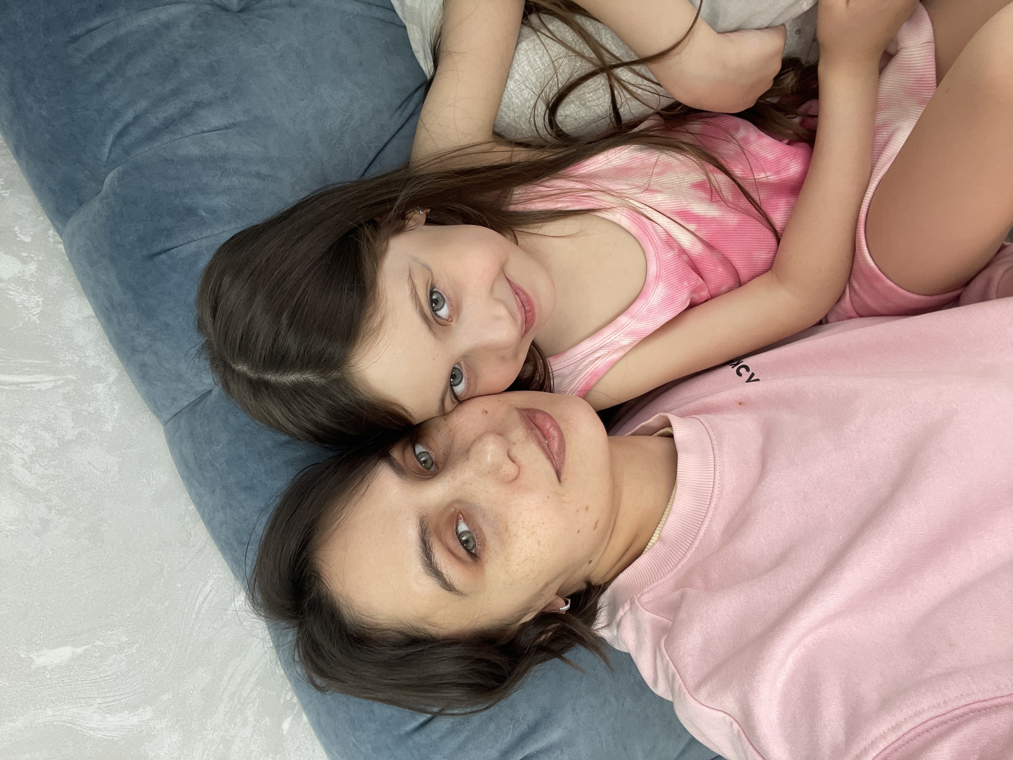 Two people, likely a mother and daughter, are lying on a blue upholstered surface. The daughter, a young girl with long brown hair and blue eyes, is wearing a pink and white tie-dye tank top. The mother, with short brown hair, is wearing a pink sweatshirt. They are positioned close together, face up, looking towards the camera. The location is Izhevsk, Russia.
