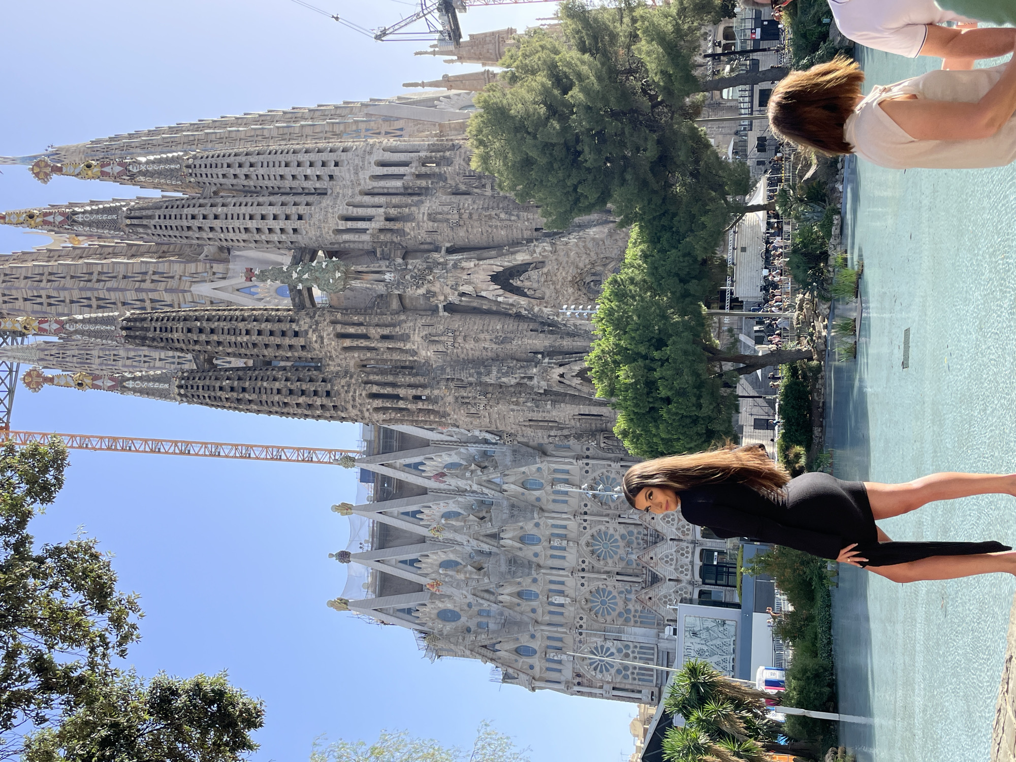 In Barcelona, Spain, in front of the Sagrada Familia, a woman in a black dress stands by a pool of water. She is facing away from the camera, with her head turned to look back over her shoulder. The Sagrada Familia, a large and ornate basilica, is visible in the background, with construction cranes present. Trees and other greenery surround the pool, and other people are seen in the distance. The sky is clear and blue.