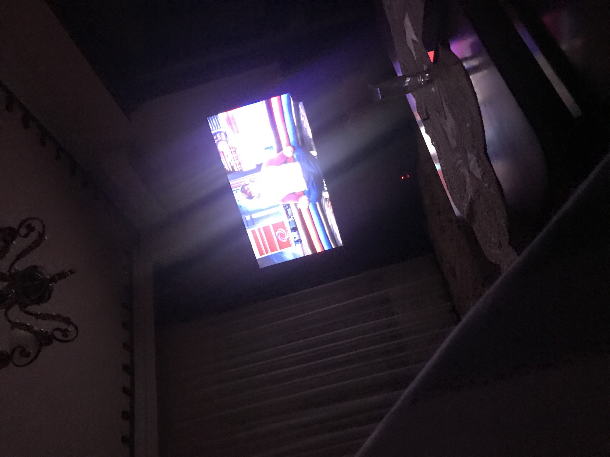 The image shows an interior view of a building, likely a church or other historical structure. A television screen is visible, displaying an image, possibly a photograph or video, with people and objects depicted in bright colors. The architecture features dark, aged materials, including stone or plaster walls and a series of steps. An ornate metal chandelier is partially visible in the lower left corner. The light from the television screen illuminates parts of the surrounding area, creating a contrast between the bright screen and the dimly lit architecture. The scene is captured from a low angle, looking upwards towards the television and the architectural details. The city and country are Yerevan, Armenia.