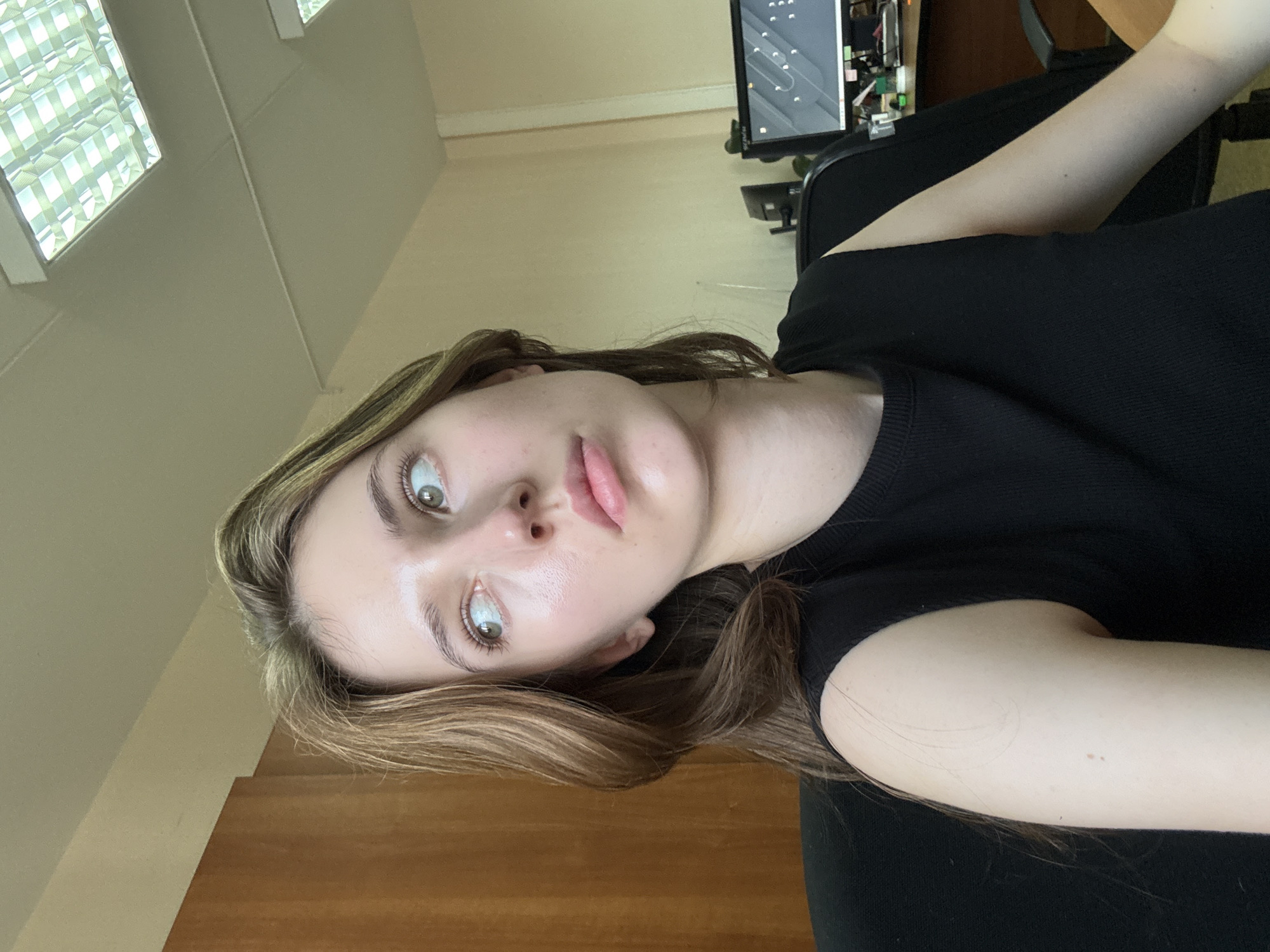 In a medium shot, a young woman with shoulder-length brown hair and light green eyes looks up and to her right. She is wearing a black sleeveless top. Her skin appears to have a subtle sheen. The background consists of an office environment. To her left is a light brown wooden cabinet. To her right, a desk is visible with a computer monitor displaying graphics, a monitor stand, and some office supplies. Above her, fluorescent ceiling lights are visible. The scene is likely an office in the city of Perm, Russia.