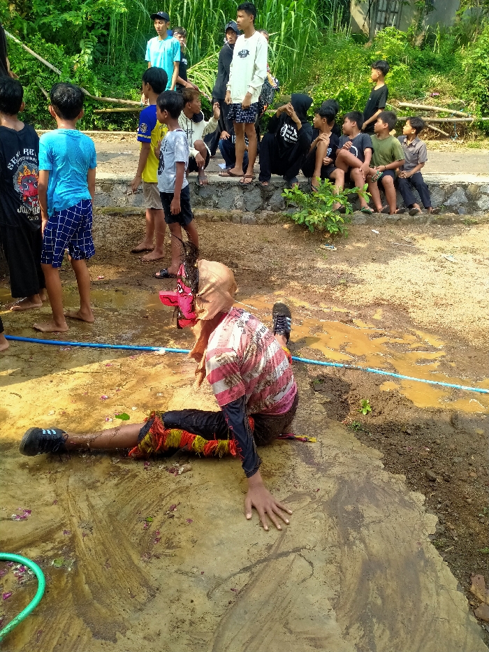 The image depicts an outdoor scene in Gebyog, Indonesia, focusing on a person partially submerged in mud during what appears to be a traditional performance or ritual.
In the foreground and center, a young male is lying on a muddy, wet concrete and dirt surface, positioned on his left side with his right arm extended forward, hand flat on the ground. He wears a light pink and white striped short-sleeved shirt over a black long-sleeved shirt, black shorts with red, yellow, and green fringe-like adornments around the lower legs, and dark athletic shoes. A light brown cloth covers his head and face, with a prominent red, possibly equine-themed, mask-like object positioned near his face. The ground around him is saturated with water and mud, with scattered red petals or debris. A green hose is visible in the far left foreground, and a blue pipe or hose runs horizontally across the muddy area.
In the mid-ground, several young males stand barefoot, observing the central figure. They wear casual attire including a black graphic t-shirt, a blue t-shirt with checkered shorts, a yellow t-shirt, and a grey t-shirt.
In the background, a group of approximately ten individuals, including young males and at least one female in a black hijab, are seated on a low stone and concrete retaining wall. They are also casually dressed, wearing t-shirts, shorts, and a light-colored long-sleeved shirt. Behind them, dense green foliage, consistent with a rural or semi-rural environment, forms the backdrop. The area behind the wall shows indications of a dirt path.