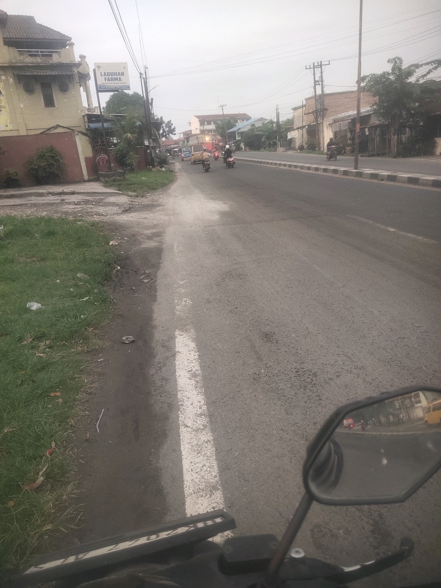 The image captures a street scene in Medan, Indonesia, likely during daylight hours with overcast conditions. A motorcycle's handlebars and side mirror are prominently featured in the foreground, indicating the photo was taken from a rider's perspective. The street itself is asphalt, marked with a white line on the right side. A grassy verge with some scattered debris runs along the left side of the road, adjacent to a building.
To the left, a building with a brownish-red lower section and a light yellow upper section is visible. A sign for "LABUHAN FARMA" is attached to a pole in front of this building, indicating a pharmacy. Further down the street, other buildings and structures line the road, some appearing to be commercial establishments.
Several vehicles are present on the road, including motorcycles and at least one van. Some of these vehicles are in motion, suggesting the flow of traffic. The overall environment appears to be a typical urban street in Indonesia, with a mix of residential and commercial buildings and a consistent stream of motorcycle traffic. Power lines and poles are also visible, common features in urban Indonesian landscapes. The presence of a yellow van further suggests a commercial area. The scene is one of everyday street activity.