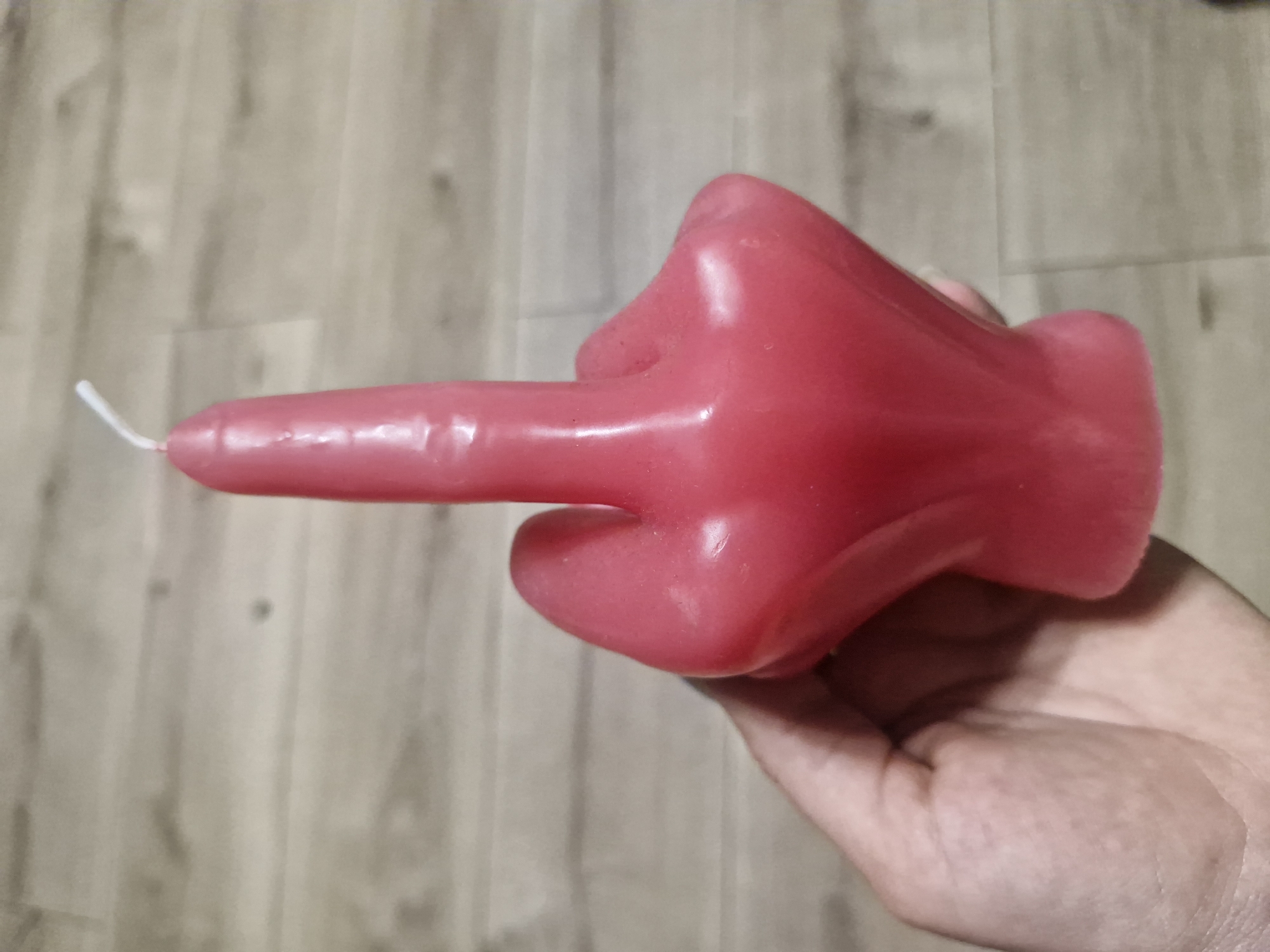 A hand is holding a red, hand-shaped candle that is giving the middle finger. The candle is positioned on a light brown wood floor. There are no indications of the location being Plovdiv, Bulgaria.