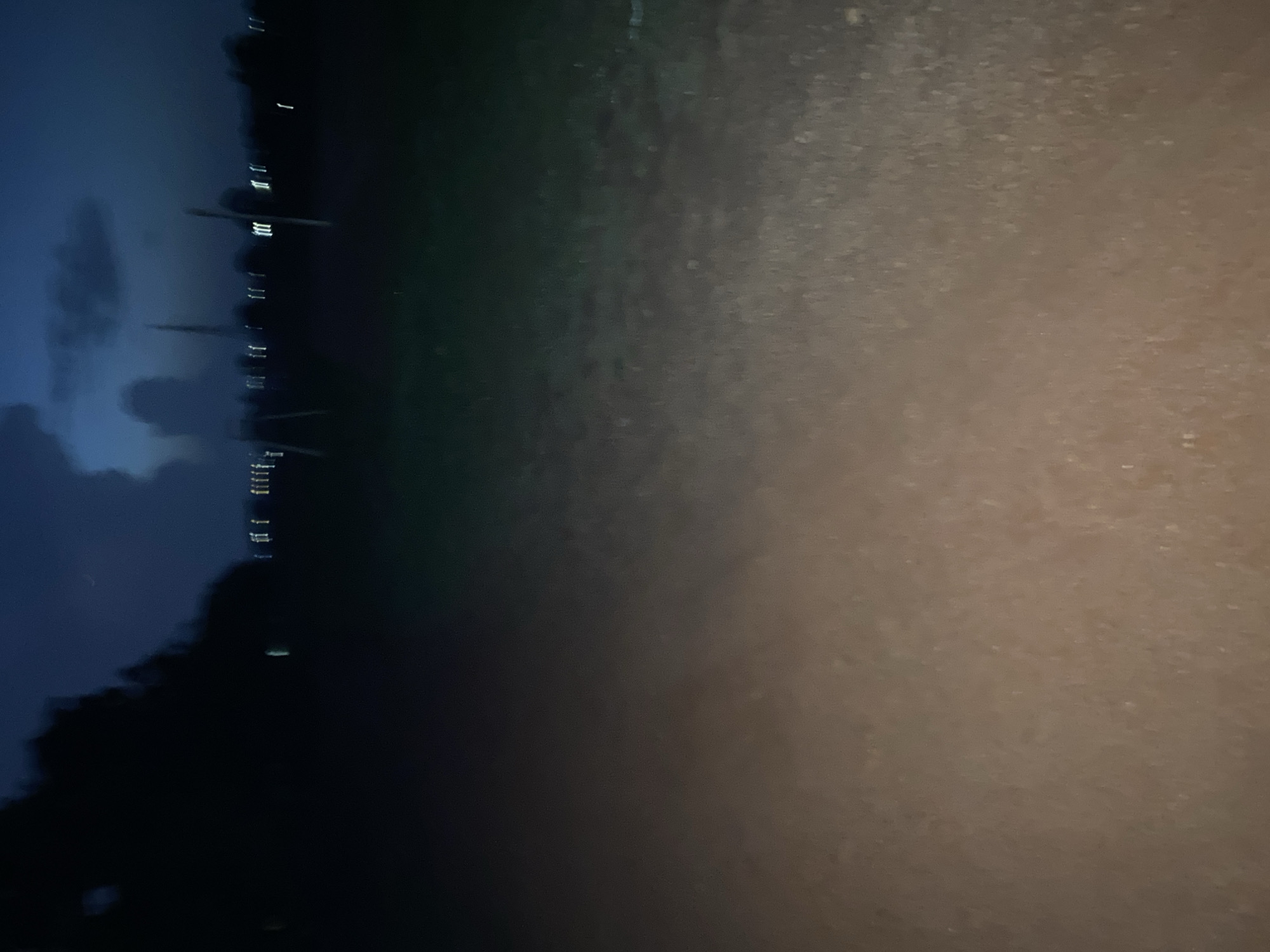 The media file presents an abstract, horizontally composed outdoor scene captured at night or during deep twilight. The left side of the image depicts a dark blue sky with subtle, darker cloud formations, merging into what appears to be a body of water. This water surface is dark blue to black, displaying numerous vertical, slightly blurred reflections of bright white artificial lights, likely originating from a distant shore or structures across the water. The reflections are streaky, suggesting either gentle water movement or a photographic long exposure.
A distinct horizontal line separates this illuminated aquatic and sky area from the right portion of the image. The right side reveals a textured, muted brown and dark grey surface, which could be foreground ground, a path, or a wall. This surface has a speckled, granular appearance and shows a gradual lightening in tone from left to right across the frame.
No identifiable people, specific objects, or discernible activities are visible, lending an abstract quality to the scene. The overall impression is one of quietude or mystery, emphasized by the darkness and artificial illumination. Given the location context of Du, Nigeria, this image likely captures a view across a local body of water, perhaps a river or lake, at night. No visible text is present in the image.
