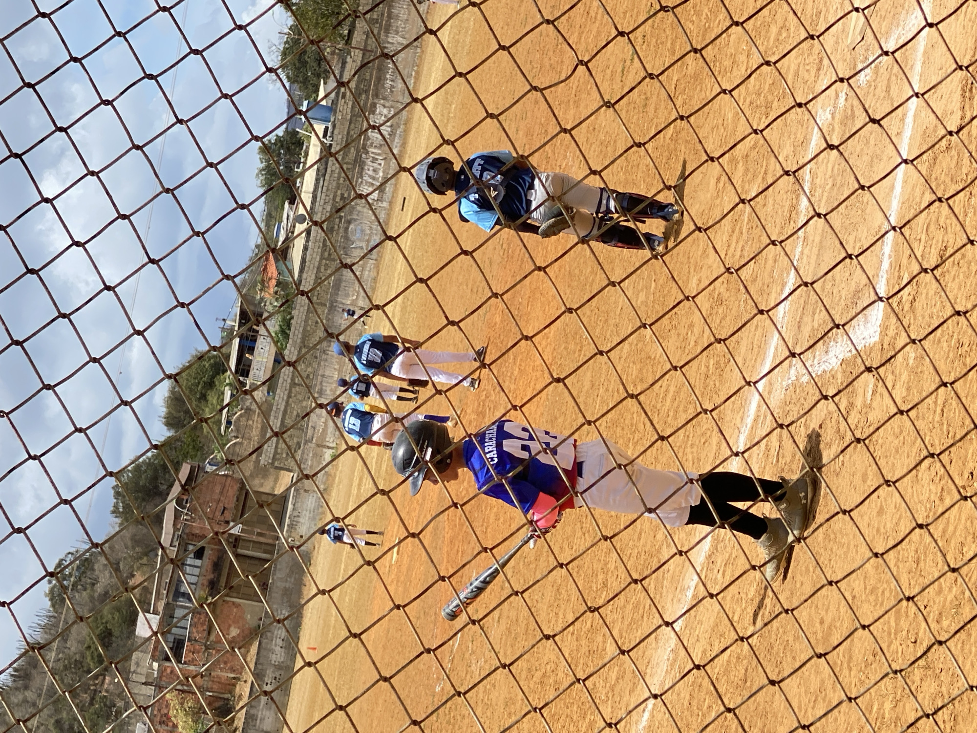 A baseball game is in progress in Altagracia, Venezuela. A player with the name "CARICHA" on the back of their jersey stands near home plate, holding a bat. Another player in catcher's gear is positioned further away, and additional players can be seen on the field. The scene is viewed through a chain-link fence. In the background are buildings and vegetation typical of the urban landscape. The playing field appears to be made of dirt or clay.